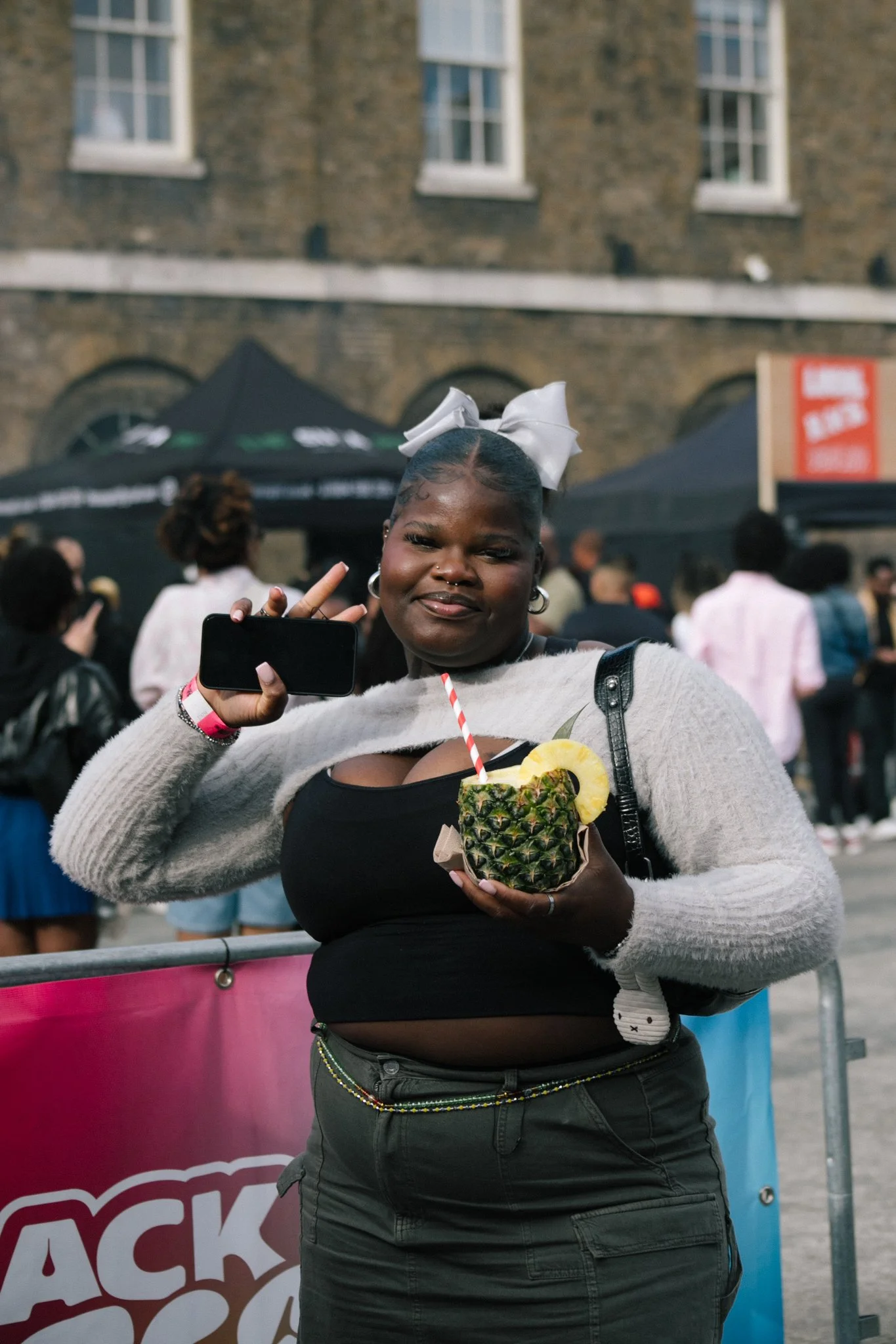 A woman holding a pineapple drink with a straw and pineapple slices, making a peace sign with her hand, at an outdoor event with black tents and a brick building in the background.