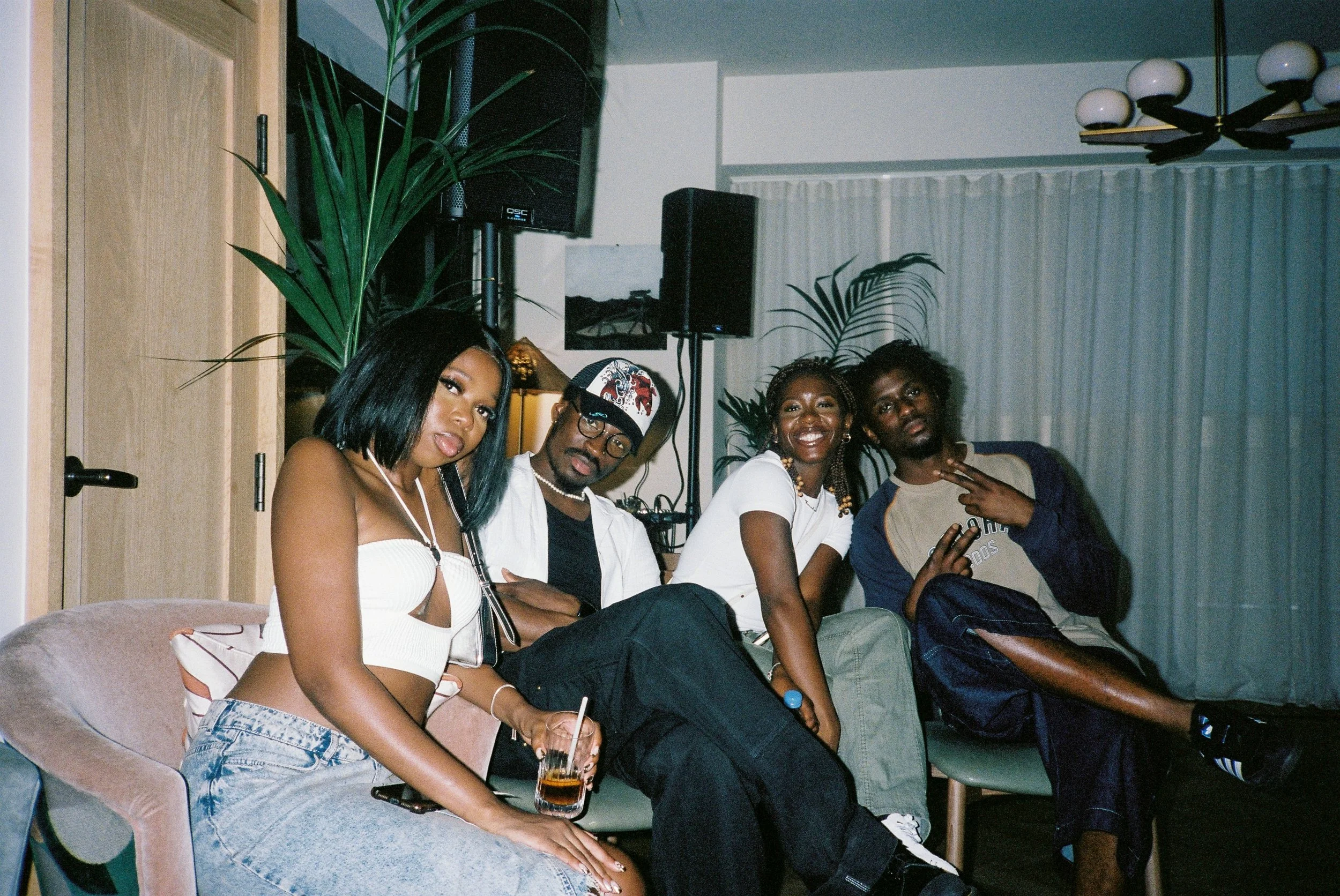 Four people sitting on a couch in a room, with two women and two men. The room has a wooden cabinet, plants, speakers, a curtain, and a ceiling fan.