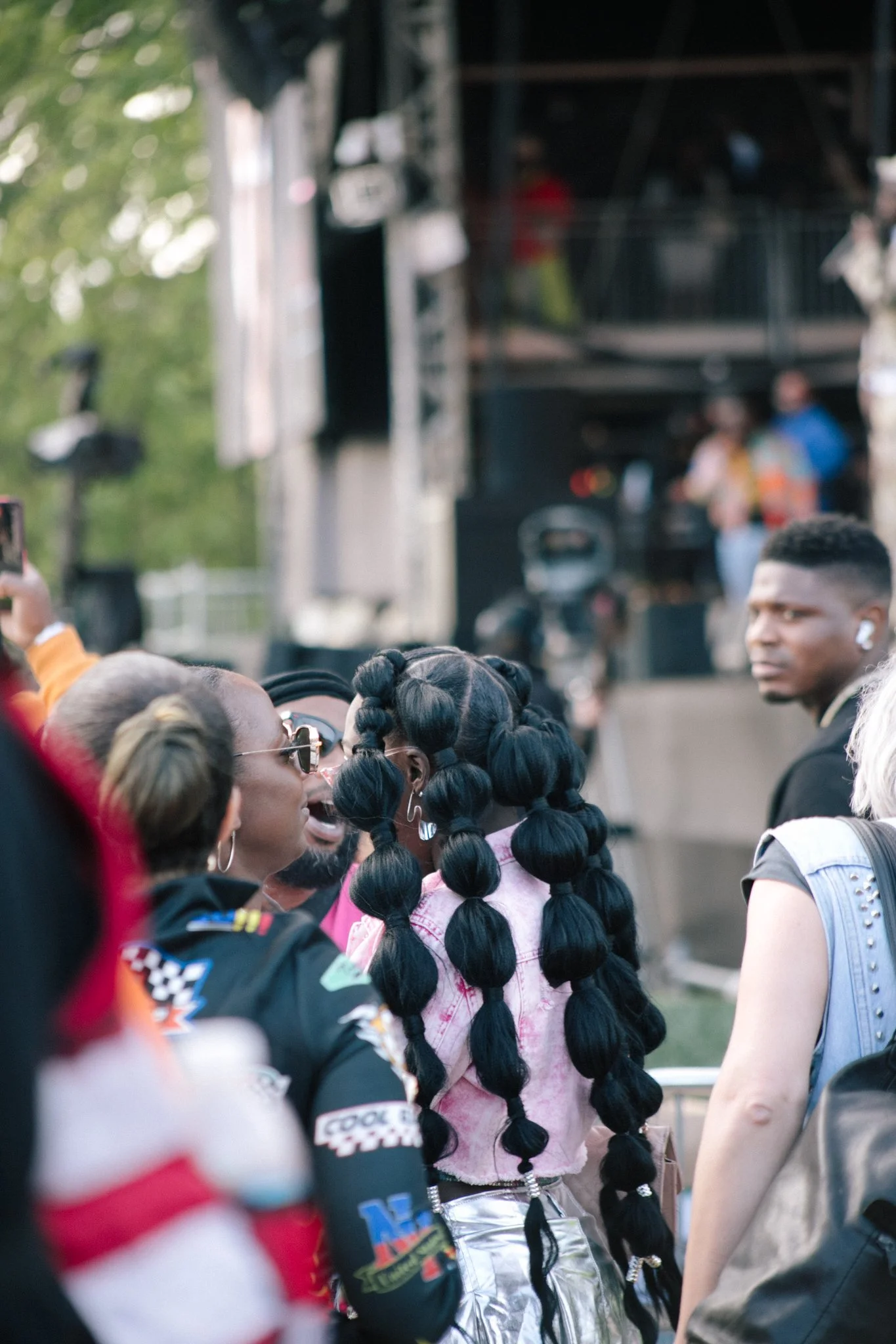 Crowd of people at an outdoor event with a stage and trees in the background. The women in focus has her back turned away from the camera and wears long bubble braids and a light pink top.