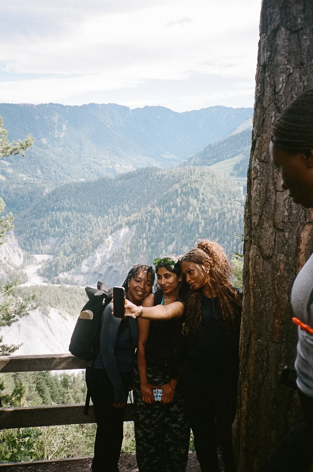Three women taking a selfie in a mountainous outdoor area with forested hills and a cloudy sky, while a woman stands partially behind a tree on the right.