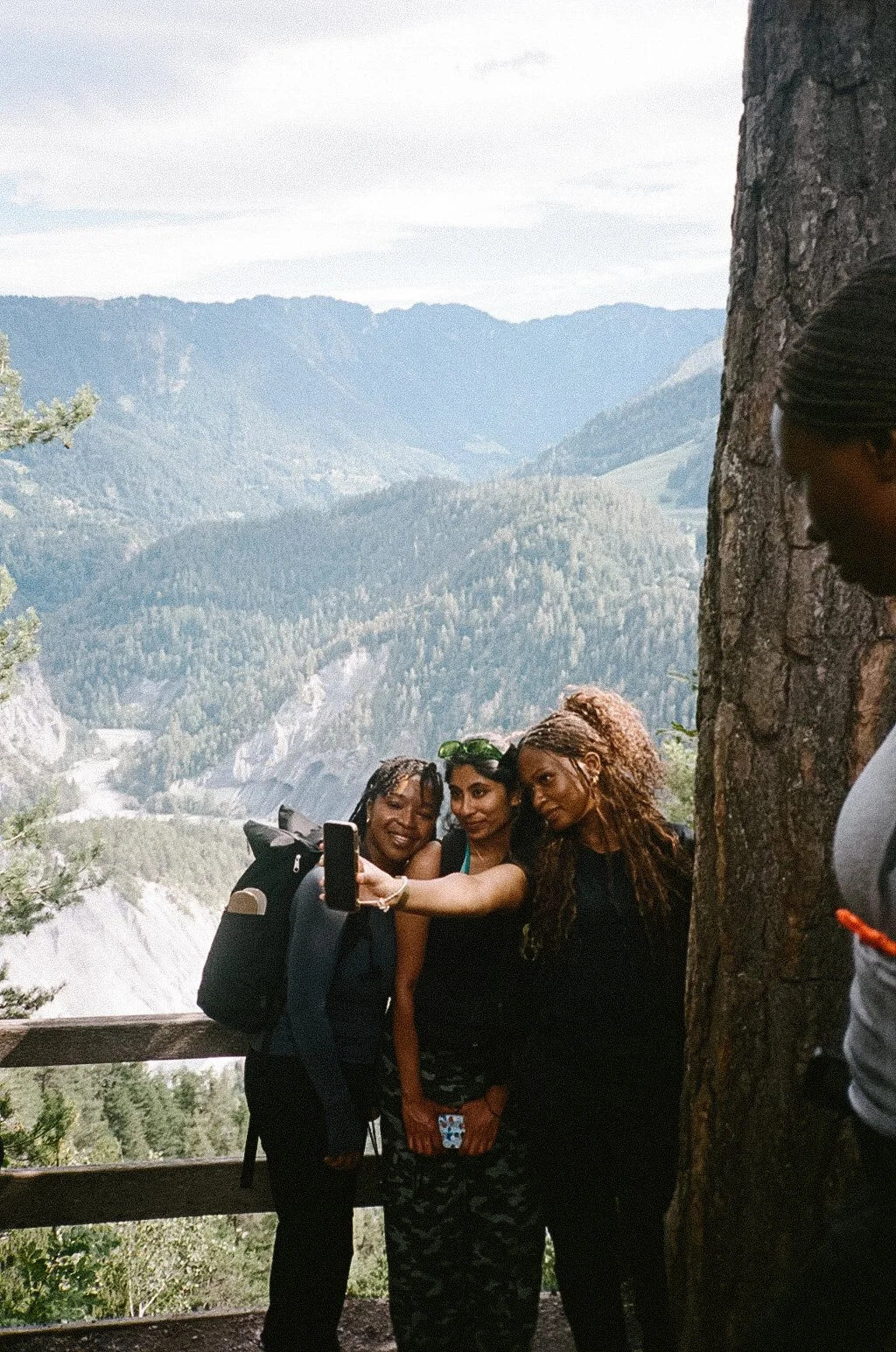 Three women taking a selfie in a mountainous area with forested hills in the background. One woman is holding a smartphone, and all are standing close to a large tree.