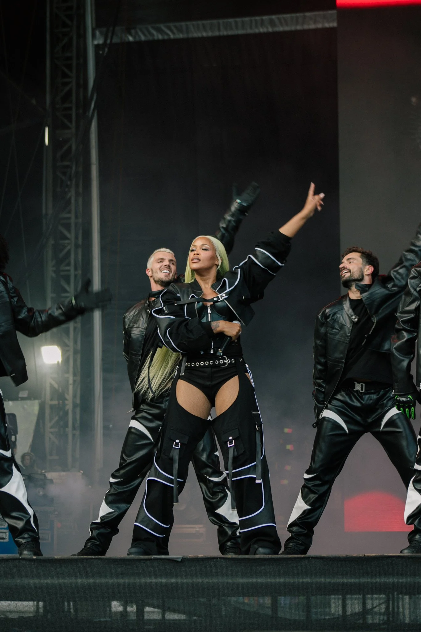 A group of five performers on stage, with female singer Eve in the center, raising her right arm and wearing a black outfit with neon accents, with male backup dancers on either side, all dressed in black with similar neon accents.
