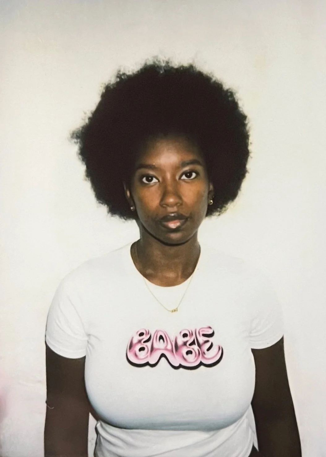 A woman with dark skin and natural afro hair wearing a white t-shirt with pink and black text that says 'BABE'. She has a gold necklace and small earrings, standing against a plain white wall.
