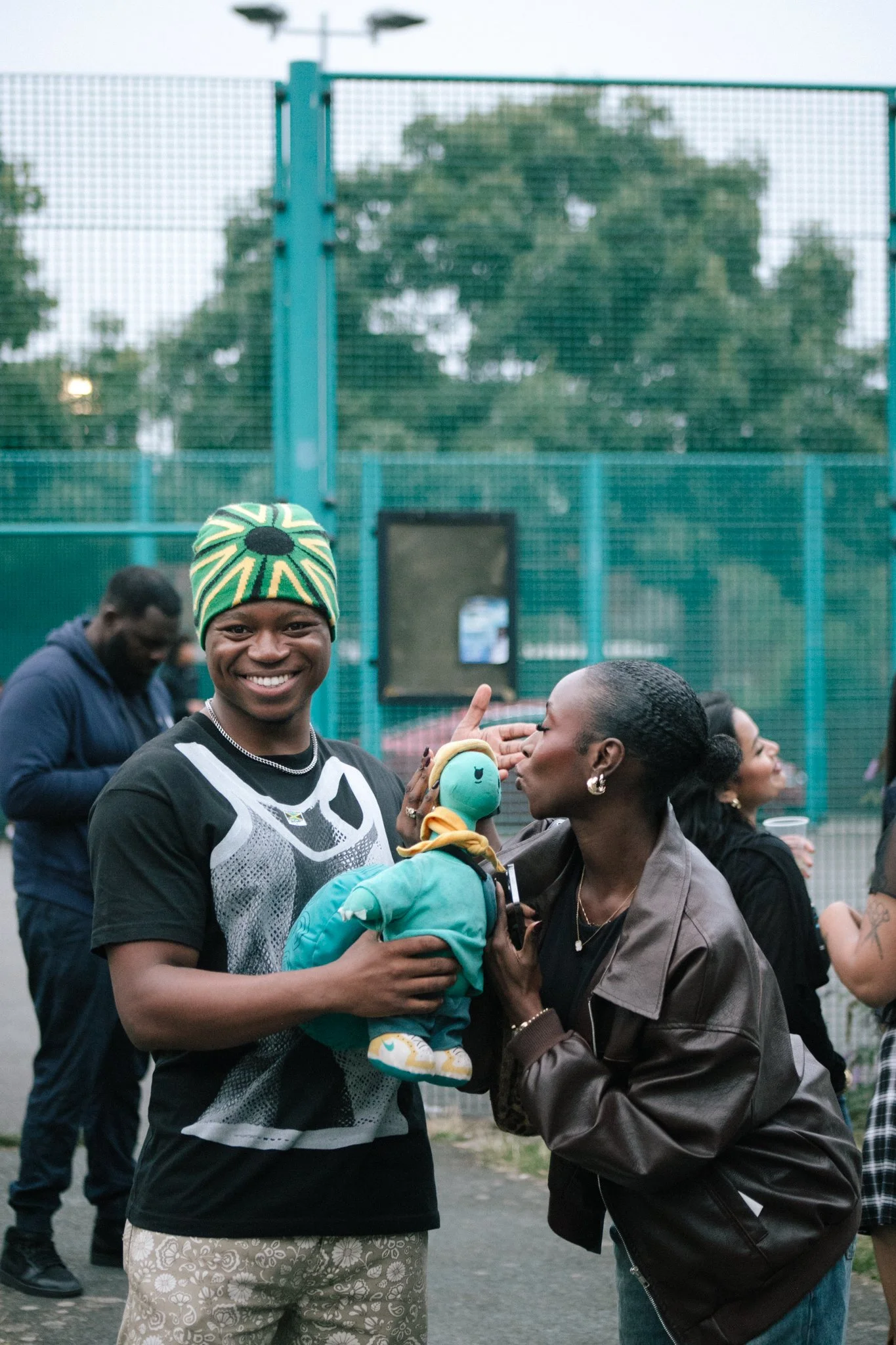A group of people, including a smiling man wearing a black and white shirt and a colorful beanie, holding a plushie in teal clothing and sneakers. A woman is giving the plushie a kiss on the cheek. 