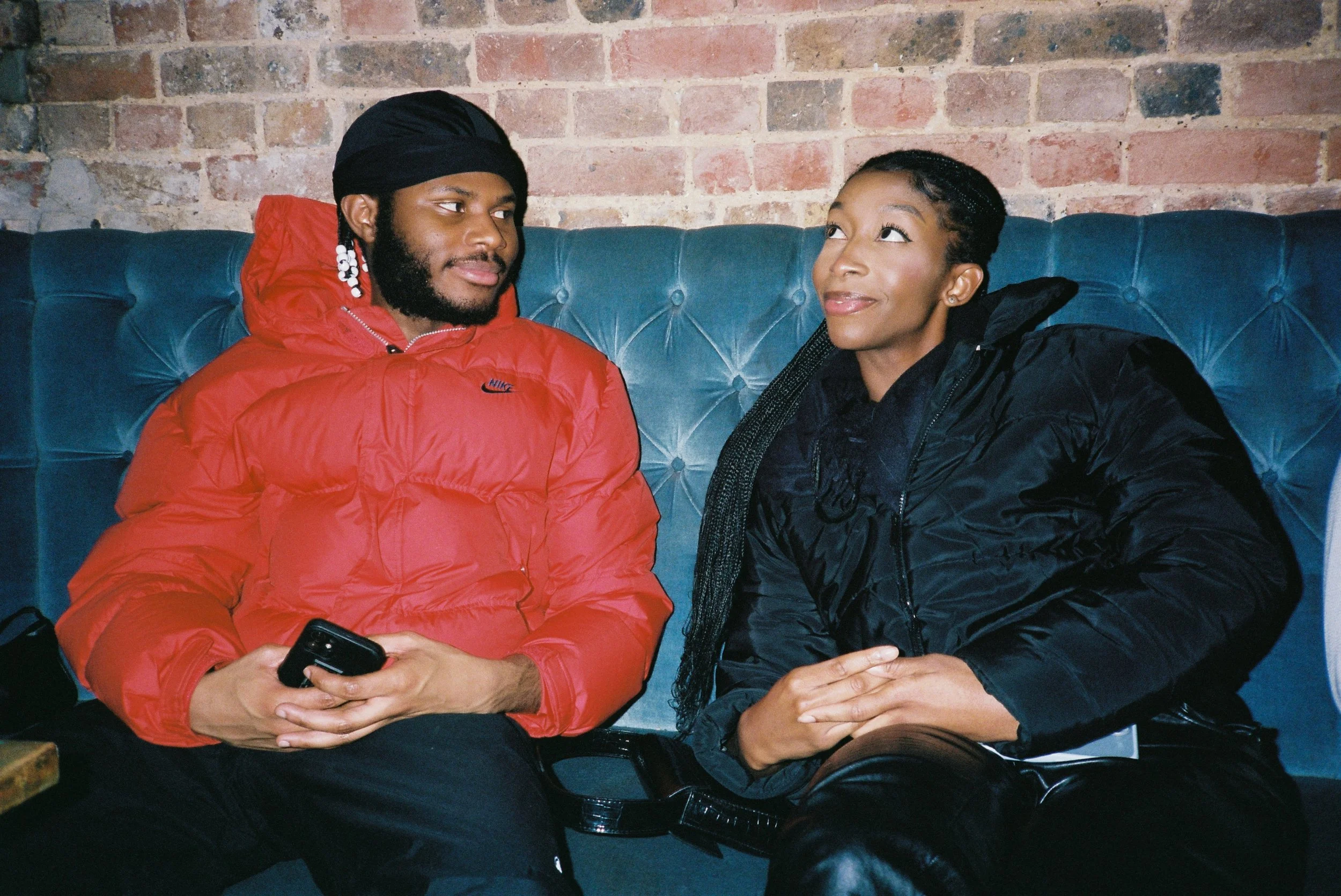 A man wearing a red jacket and black head covering sitting on a blue cushioned bench next to a woman in a black jacket, both looking at each other in a casual indoor setting with a brick wall background.