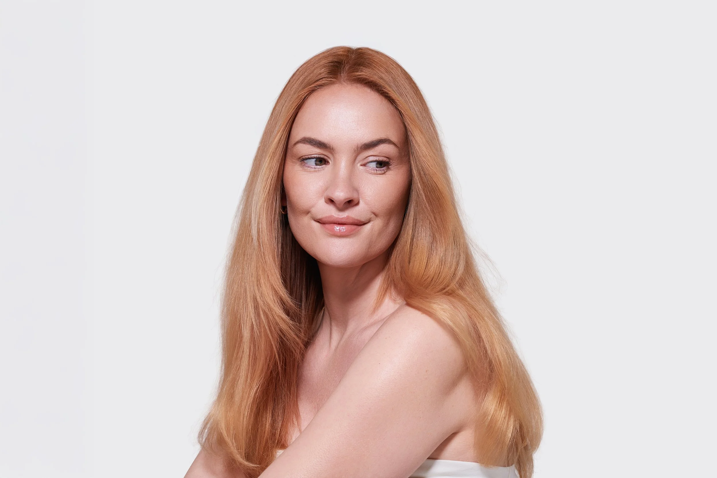 A woman with long red hair, fair skin, and minimal makeup, looking slightly downward with a subtle smile, against a plain white background.