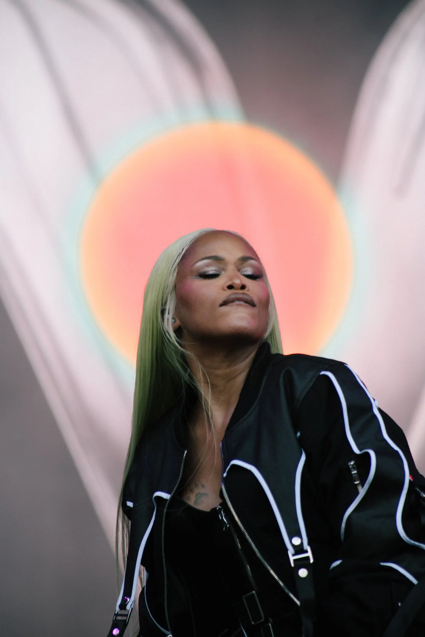 Musician Eve, wearing long green hair, makeup and a black jacket with white piping, posing in front of a multicolored circular light projection.