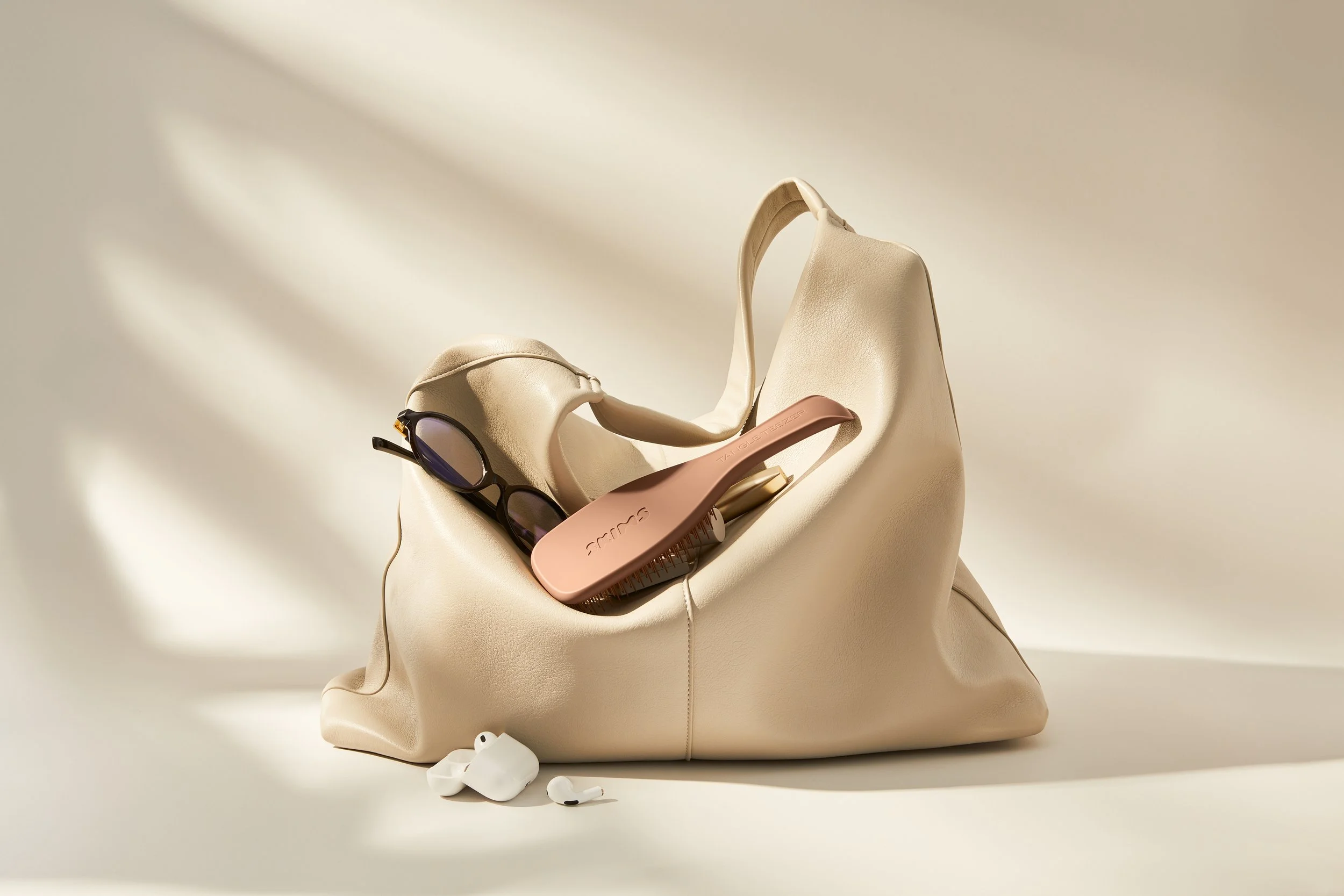 A beige slouch handbag containing reading glasses, a reddish brown hairbrush, and white wireless earbuds, all placed on a cream surface with shadow lighting.