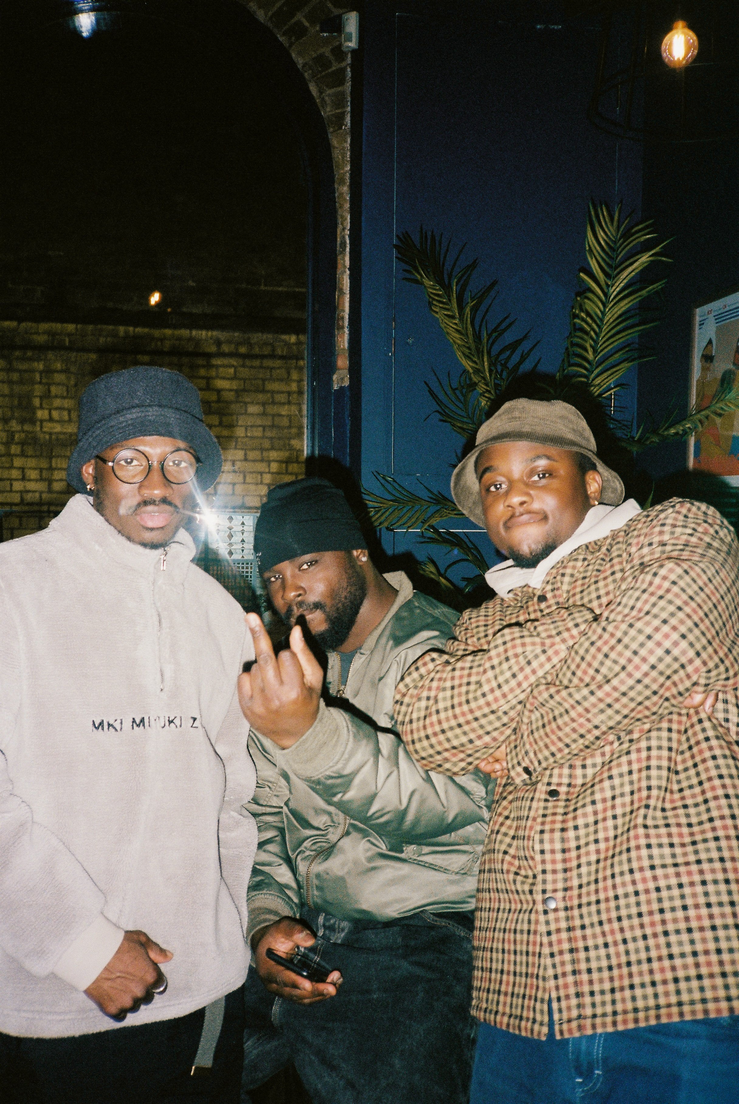 Three young men posing indoors; the man on the left wears glasses and a bucket hat, the man in the middle is making a hand gesture and holding a phone, and the man on the right wears a visor and a checkered jacket. There is a large plant and a brick 
