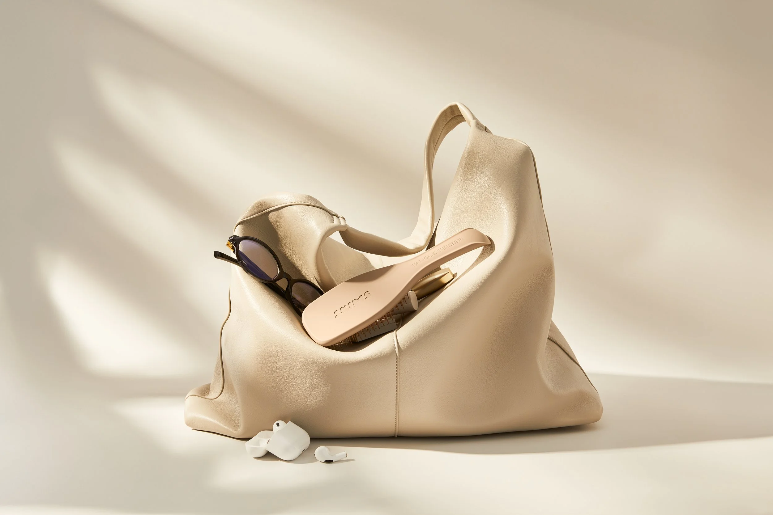 A beige slouch handbag containing reading glasses, a light brown hairbrush, and white wireless earbuds, all placed on a cream surface with shadow lighting.