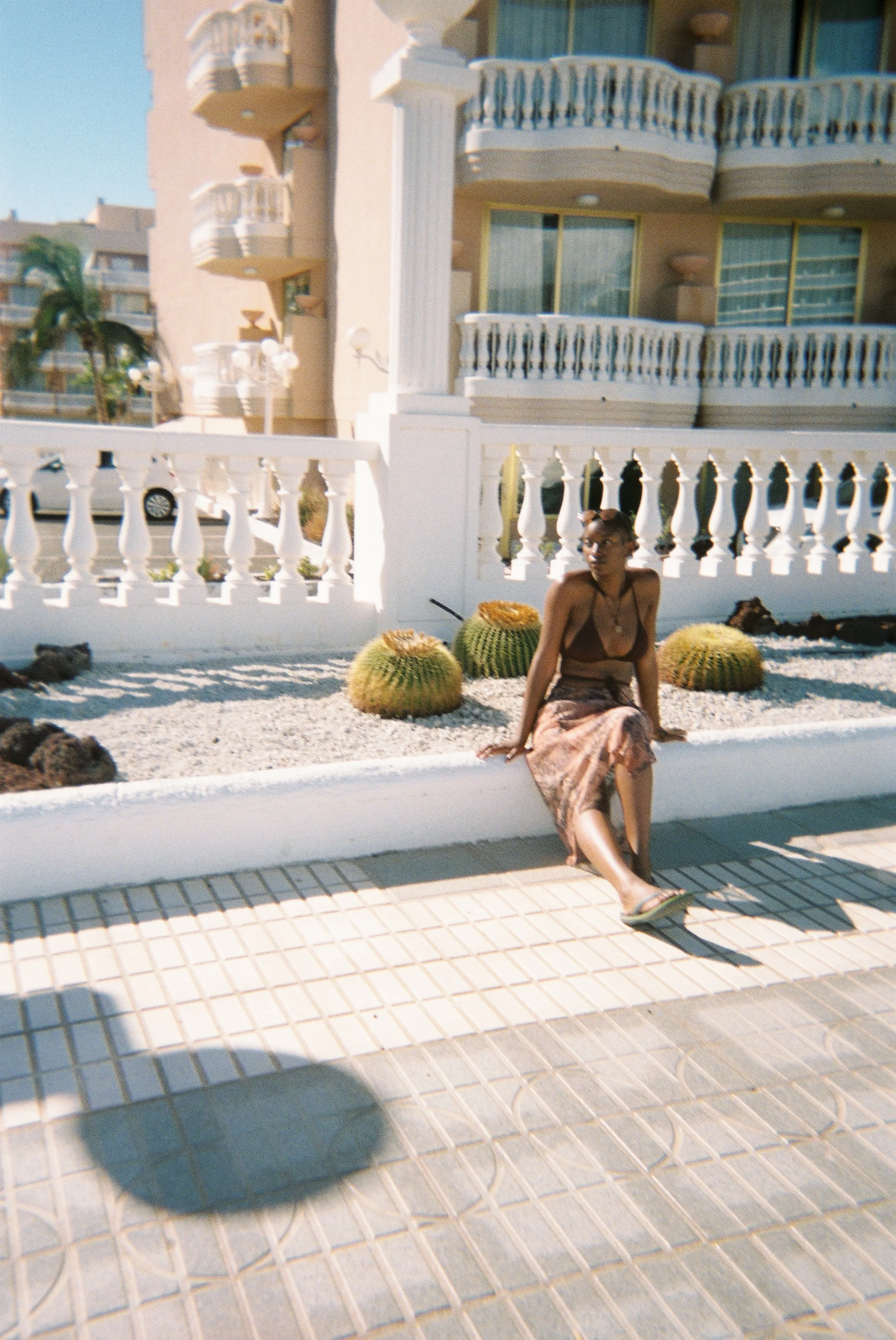 A young woman wearing a brown swim top and a patterned skirt is sitting on the edge of a sidewalk near large green cacti. The photo is taken during the daytime with clear skies and sun.