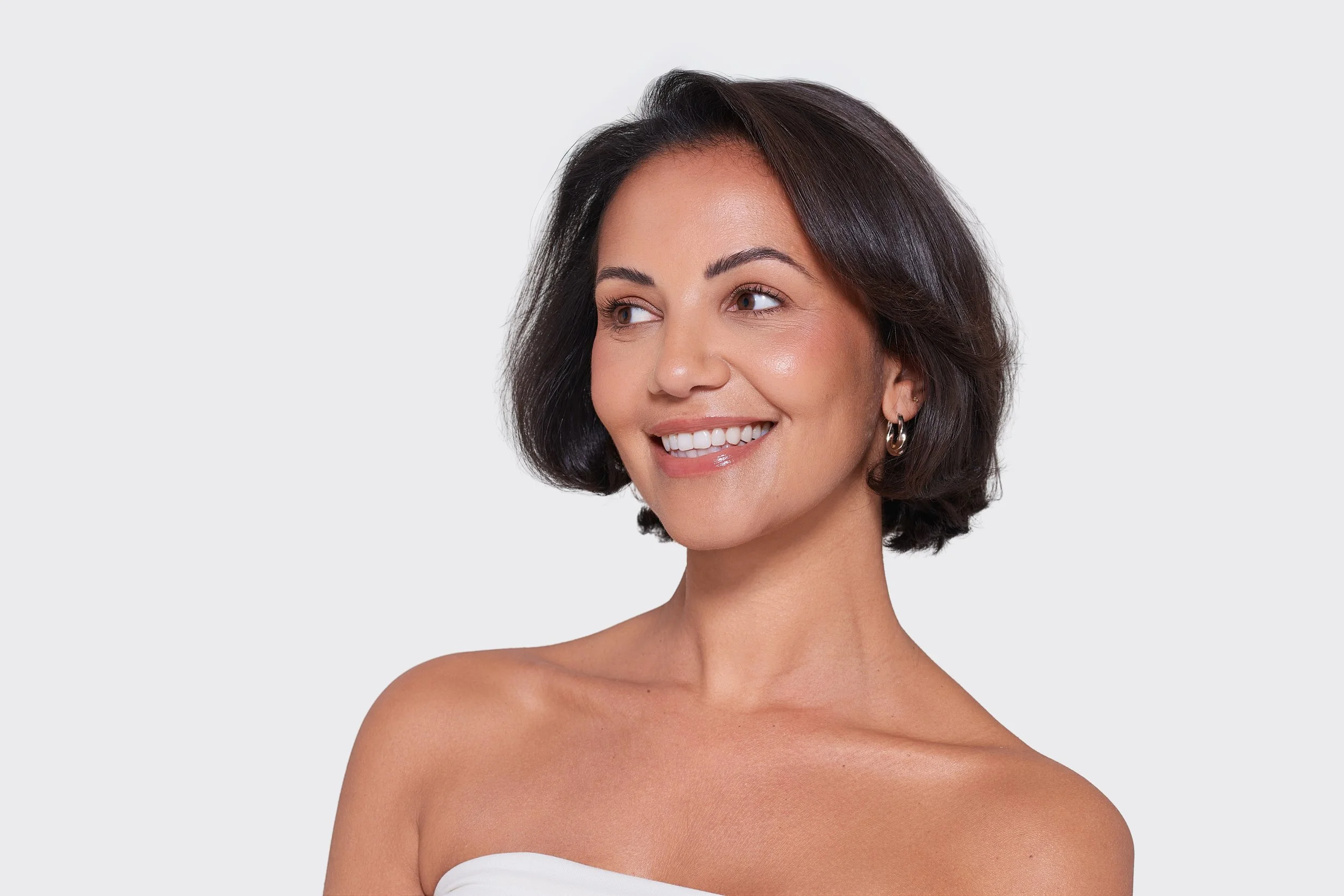 A woman with short, dark hair smiling and looking to the side, wearing hoop earrings against a plain gray background.