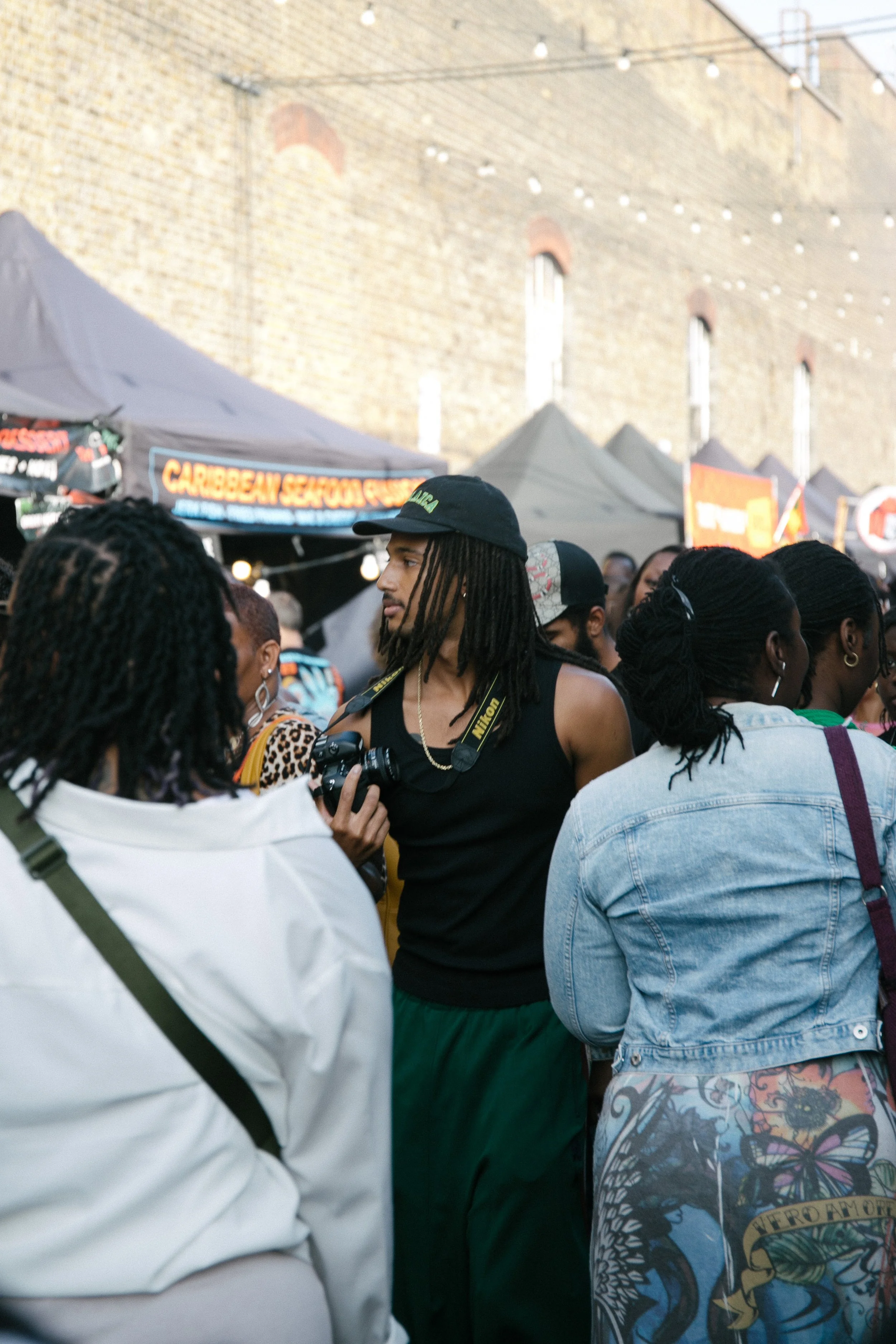 A crowd of diverse people gathered at an outdoor event with tents and string lights overhead, set against a brick building. The focus of the image is a man with locs in the center holding a camera.