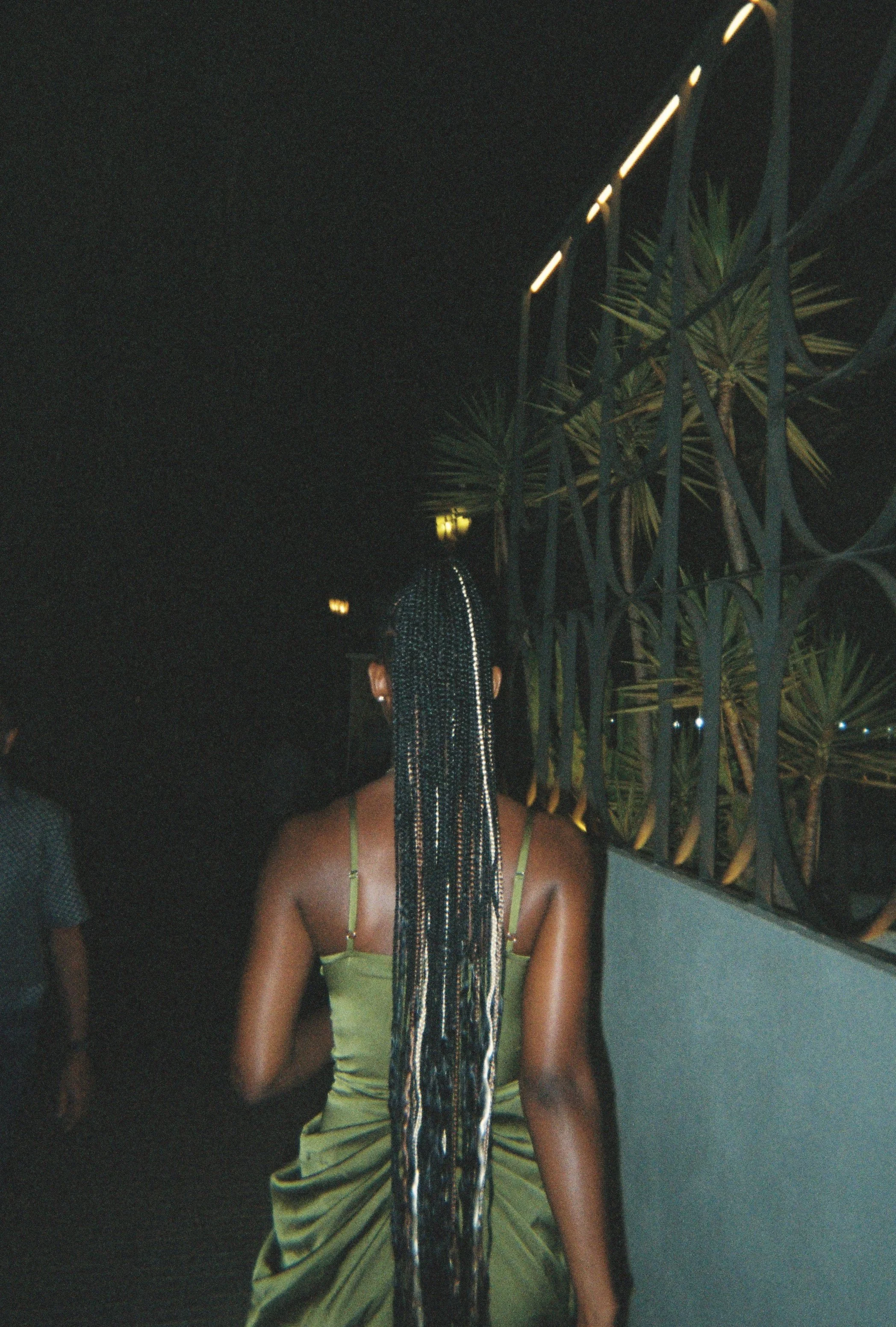 A woman with long braided hair standing in front of a decorative metal fence and plants at night, wearing a green dress with thin straps.
