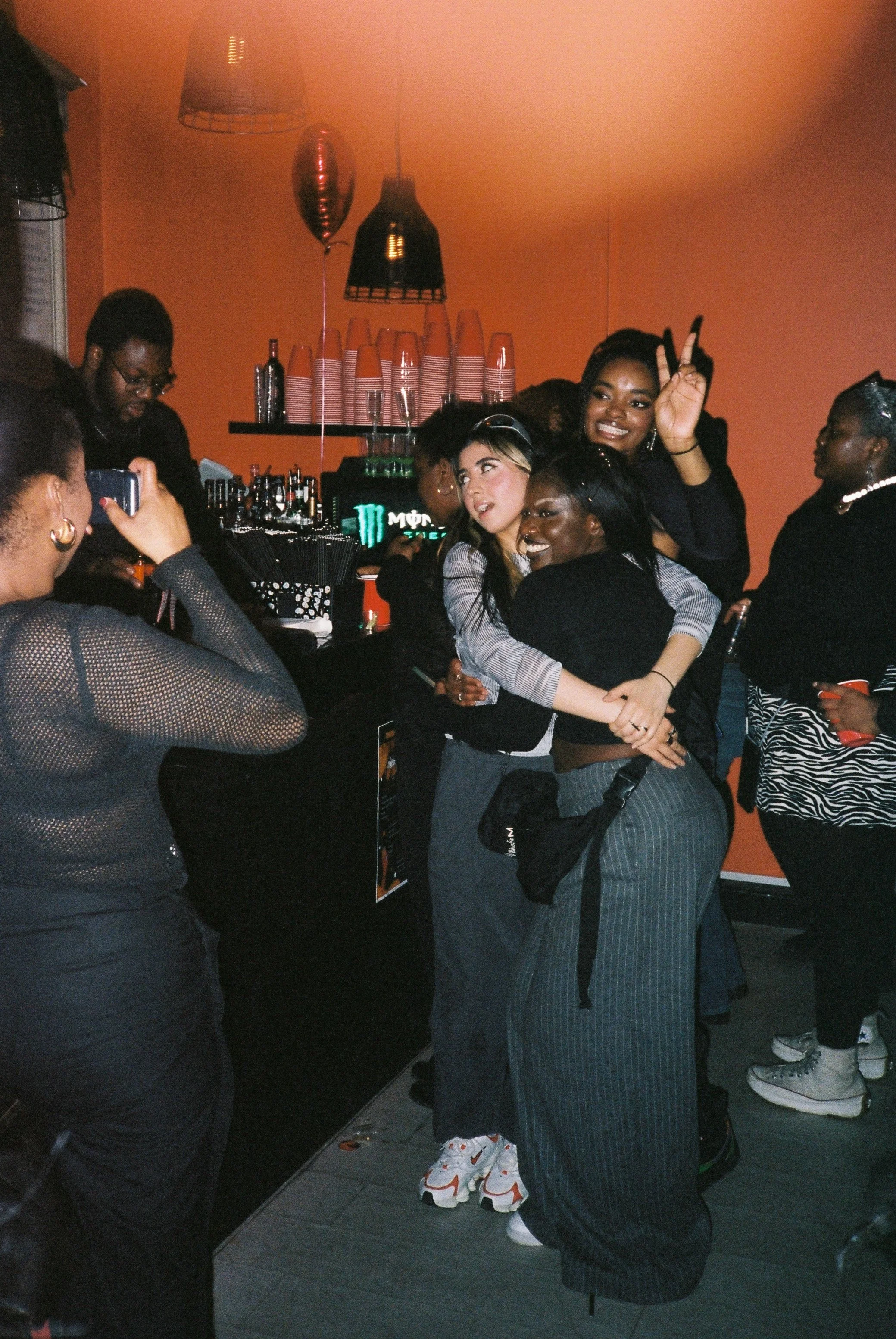 A group of young women at a lively social gathering, posing and smiling for photos inside a room with an orange wall, decorated with hanging balloons and a bar with drinks in the background.