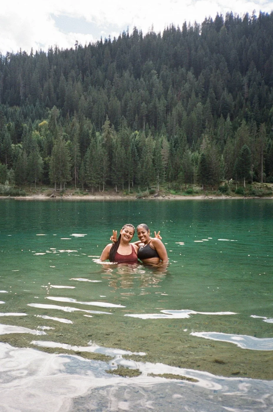 Two women smiling and making peace signs while standing in a lake surrounded by a forest and mountains.