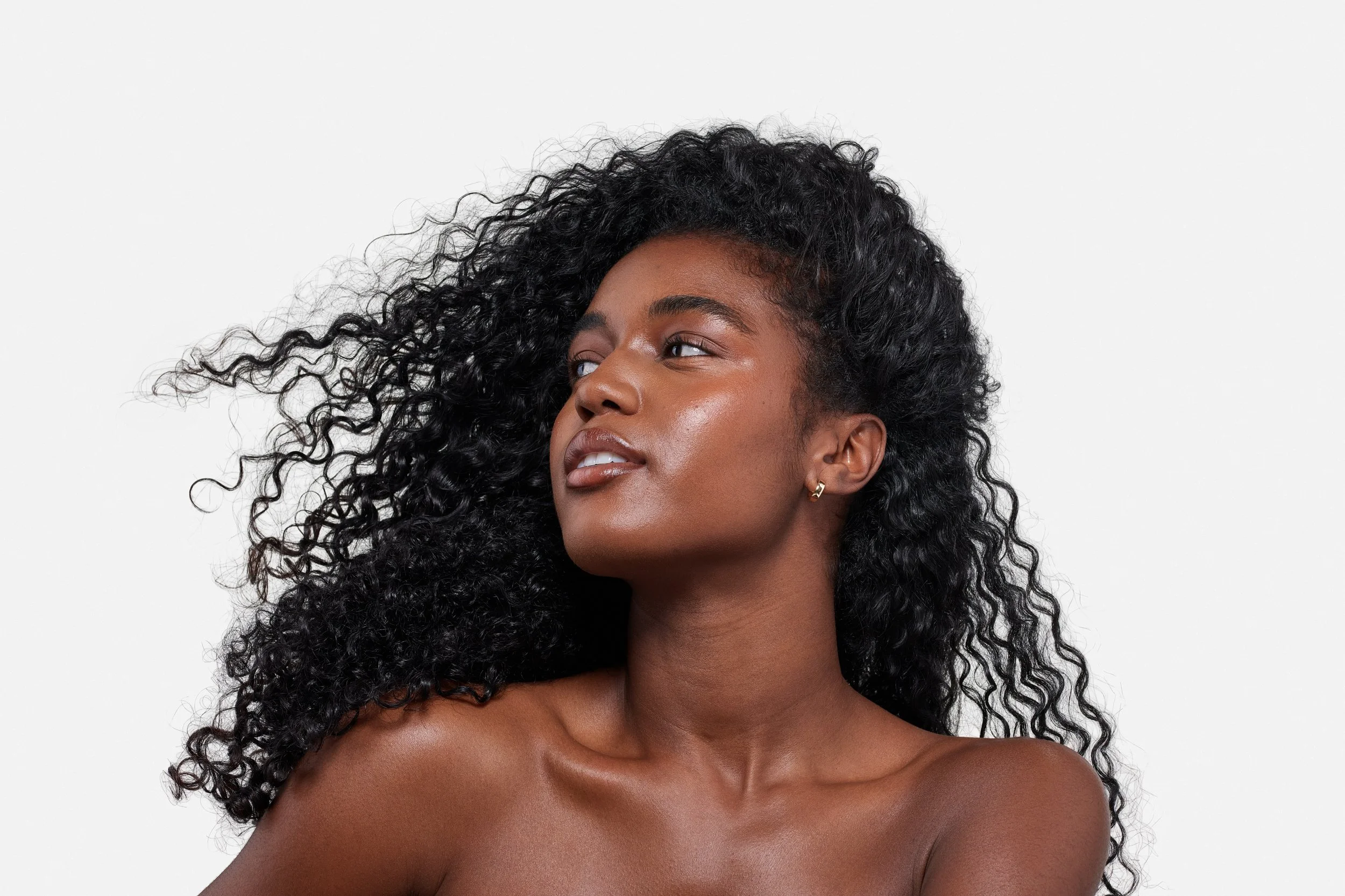 Close-up of a young woman with dark curly hair, dark skin, and gold earrings, against a plain white background.