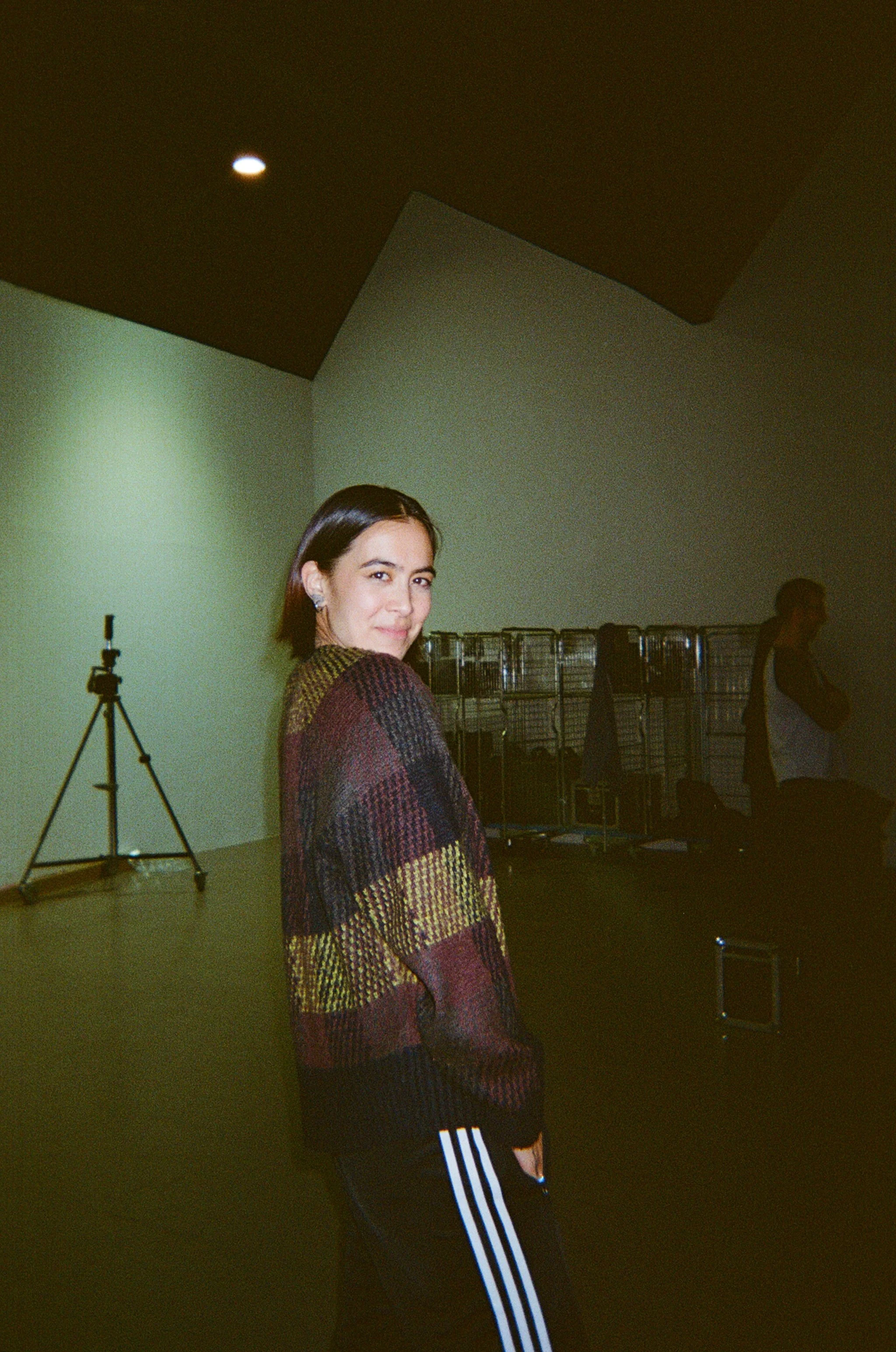 Young woman with short dark hair standing on a production set, wearing a colourful checkered sweater and black Adidas track pants.