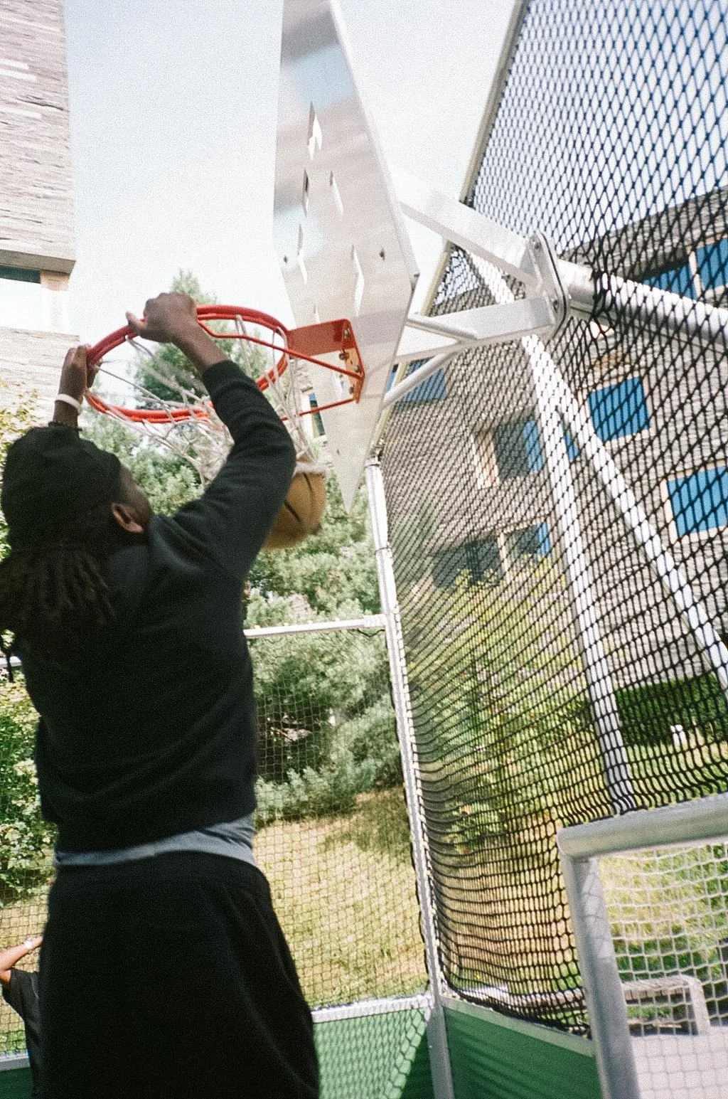 A person with dreadlocks wearing a black hoodie and black pants attempts a basketball dunk on an outdoor hoop, with a metal backboard and orange rim, during daytime. There is a wire fence and a building in the background.