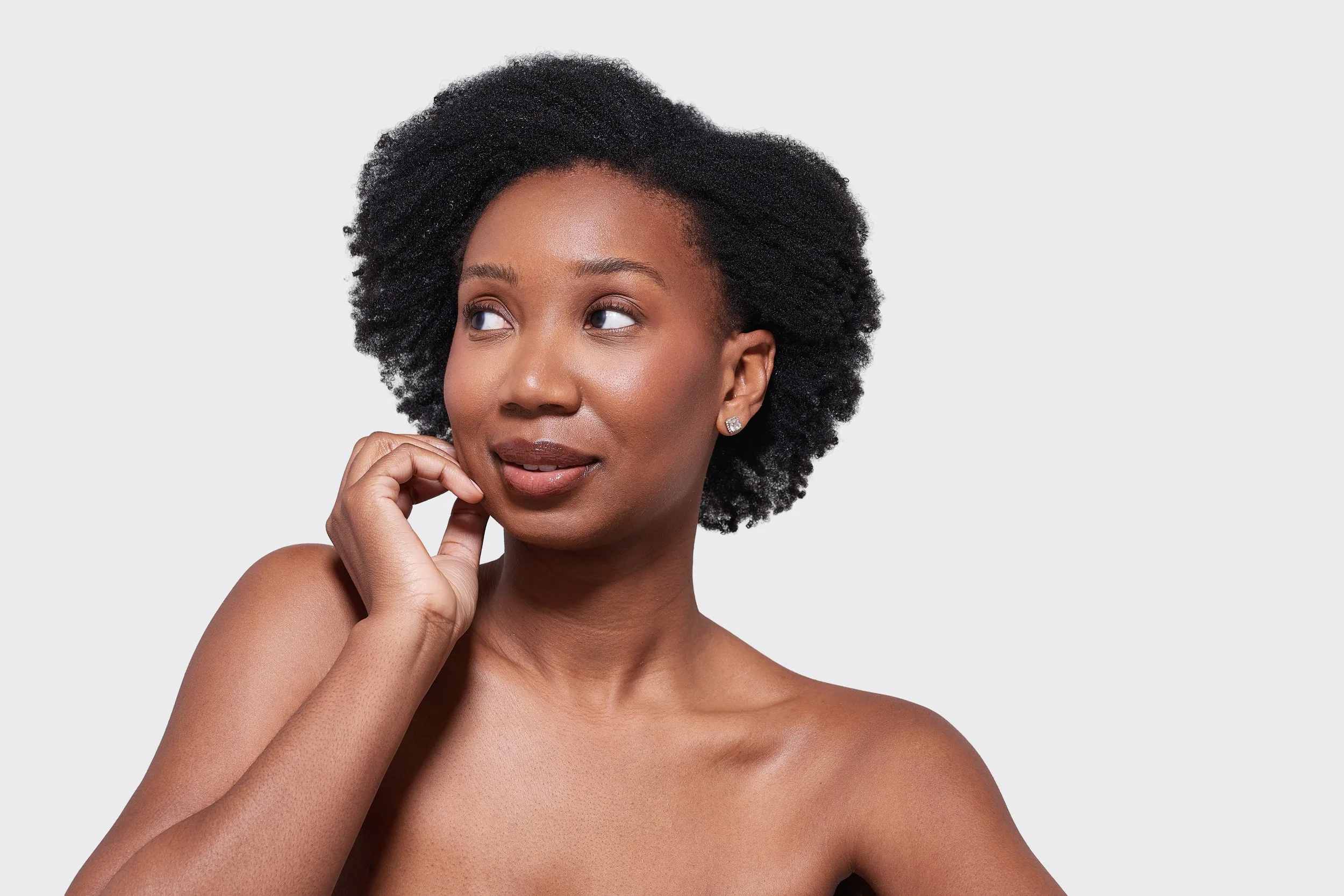 A close-up portrait of an African American woman with short, curly hair, wearing diamond earrings, looking thoughtfully to the side with her hand near her face, against a plain light gray background.