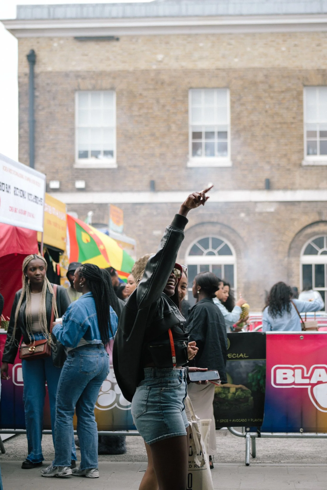 People gathered at an outdoor event, with a woman smiling and pointing upward, dressed in a black leather jacket and denim shorts, amidst a crowd with colourful stalls and banners.