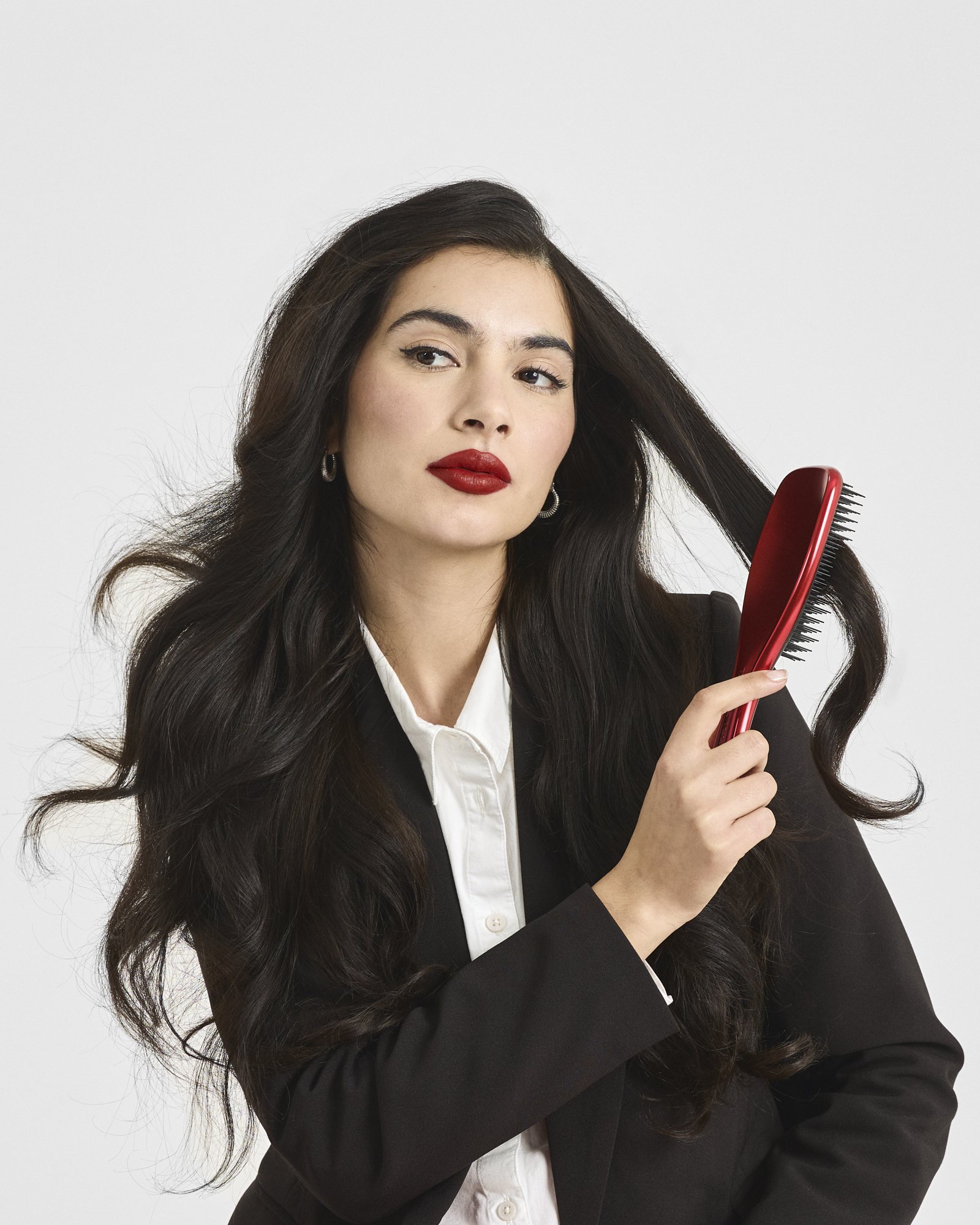 A woman with long dark hair, wearing red lipstick, a black blazer, and a white collared shirt, is looking slightly off-camera while holding a red hairbrush to her hair.