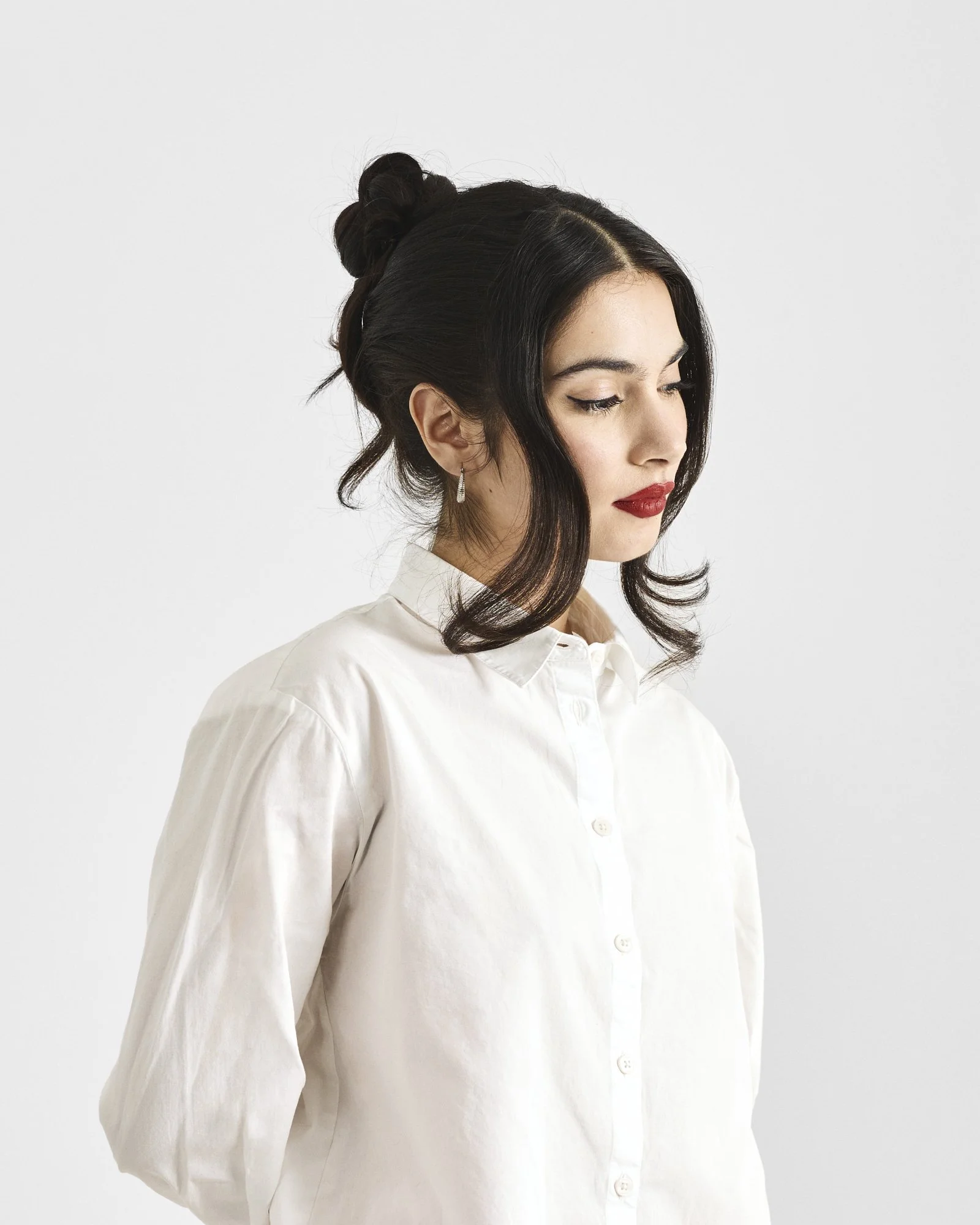 A woman with dark hair styled in an updo, wearing a white blouse and diamond drop earrings, looking down against a plain light background.