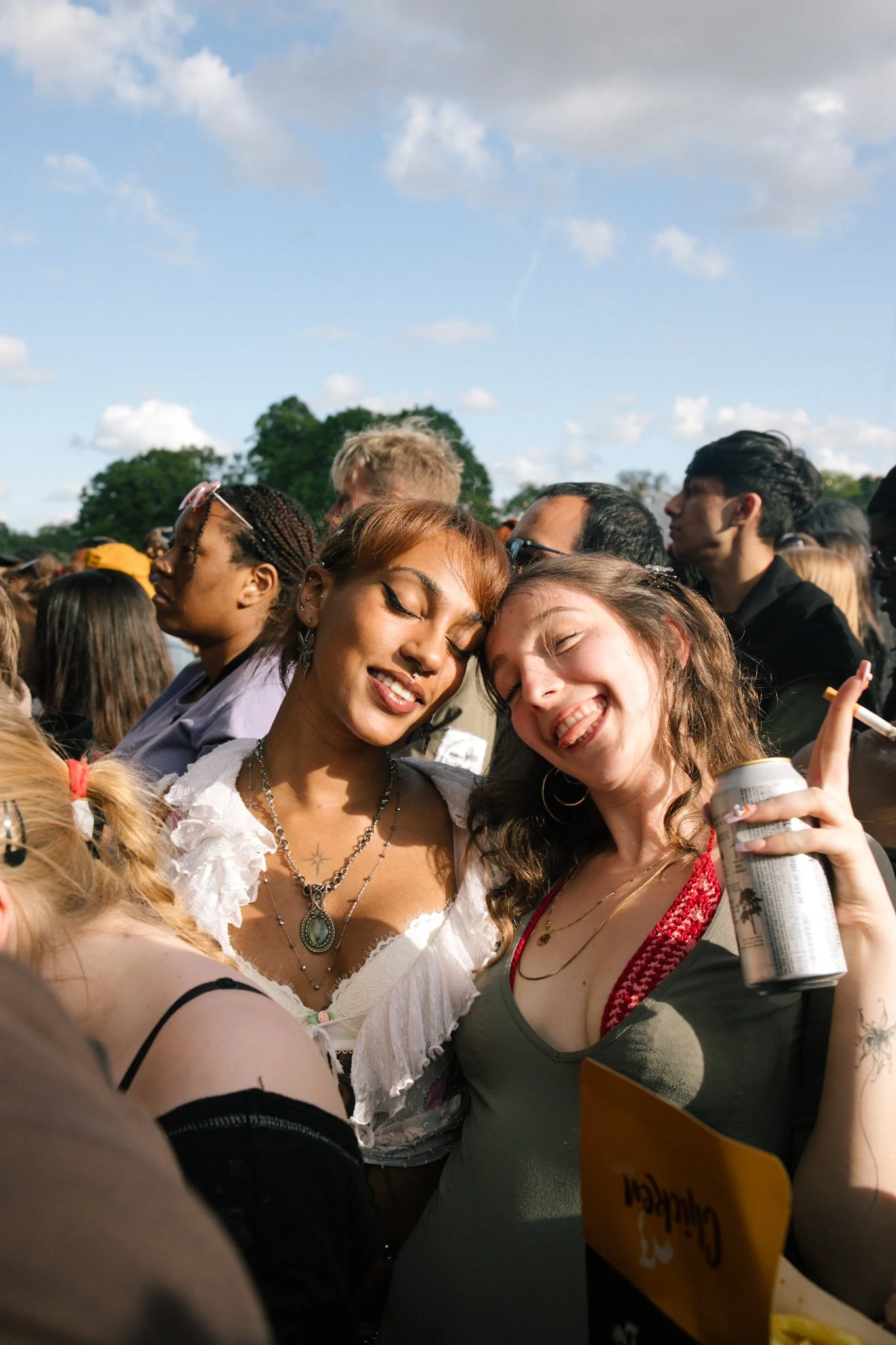 Two women hugging and smiling at a crowded outdoor event with a blue sky and clouds background.