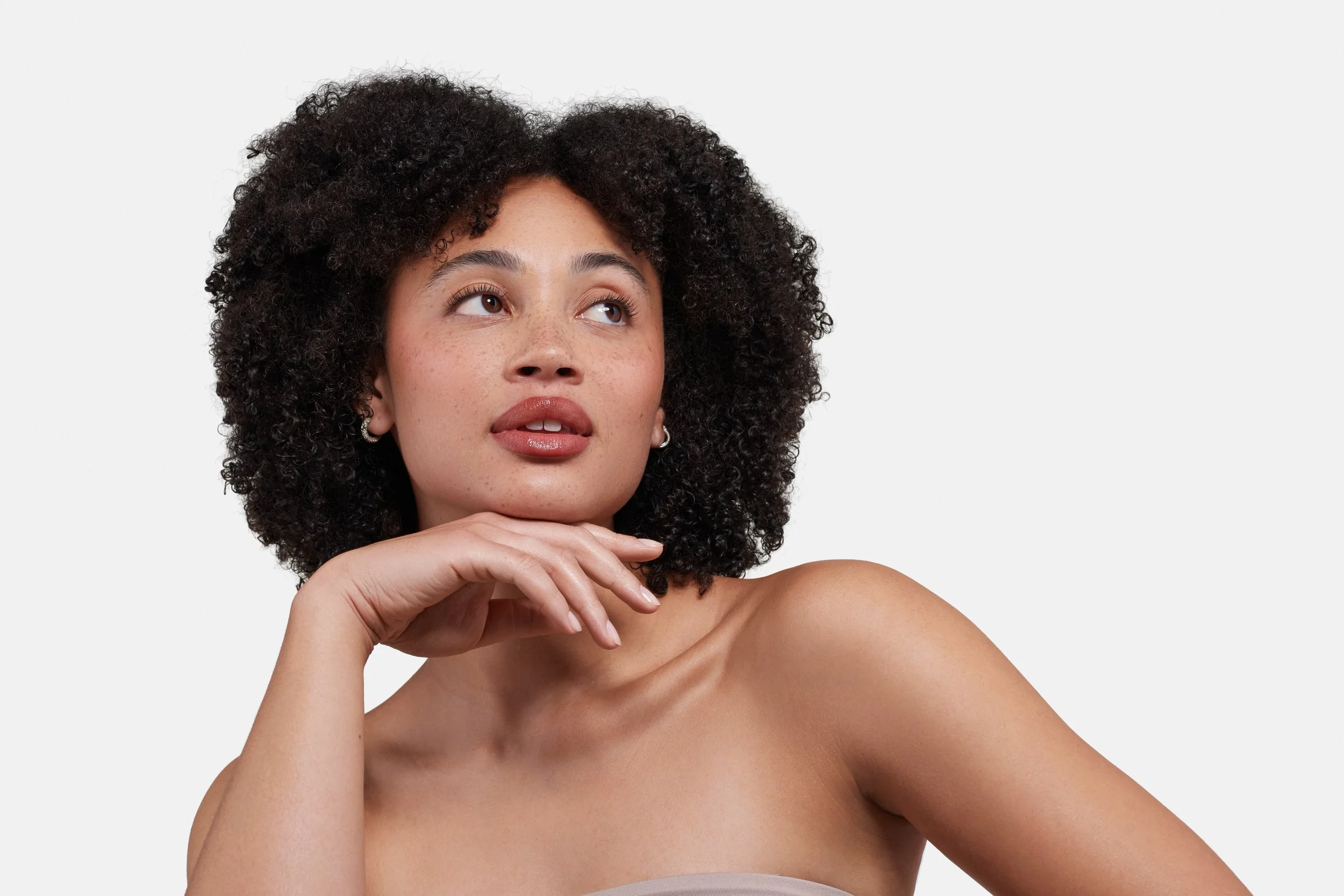 Close-up of a woman with curly hair and freckles, resting her chin on her hand, looking upward