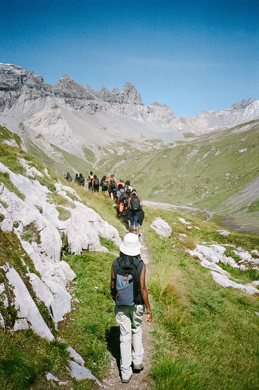 A group of hikers walking along a mountain trail with green grass and rocky terrain, under a clear blue sky, with rugged mountain peaks in the background.