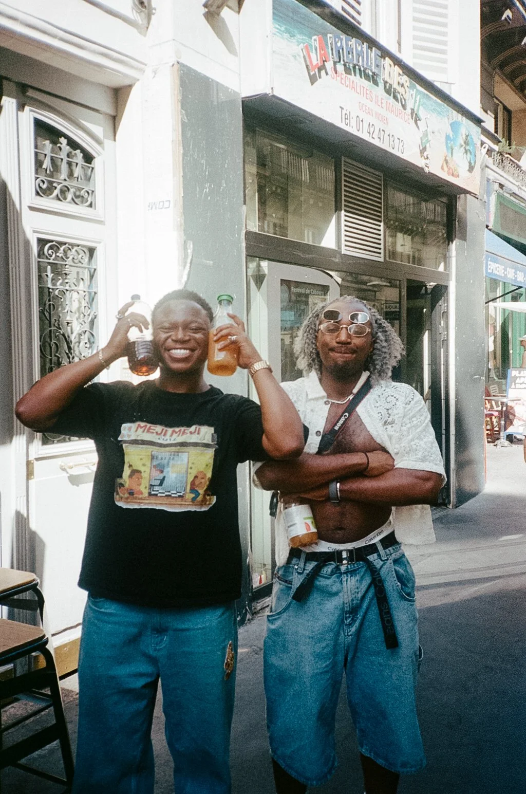 Two men standing outside a restaurant with one smiling and holding two bottles of beverages, the other with arms crossed, wearing sunglasses and a partially open shirt.