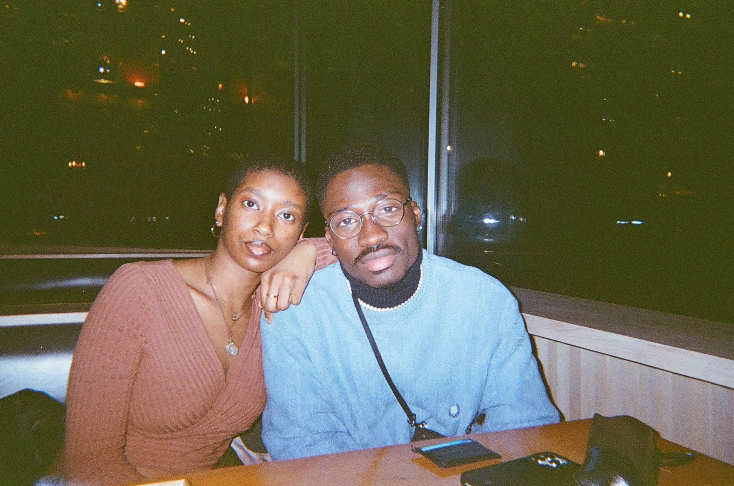 A man and woman sit close together at a table, smiling at the camera. The woman has short curly hair and wears hoop earrings, while the man wears glasses and a pearl necklace. In the background is a window with a nighttime city view.