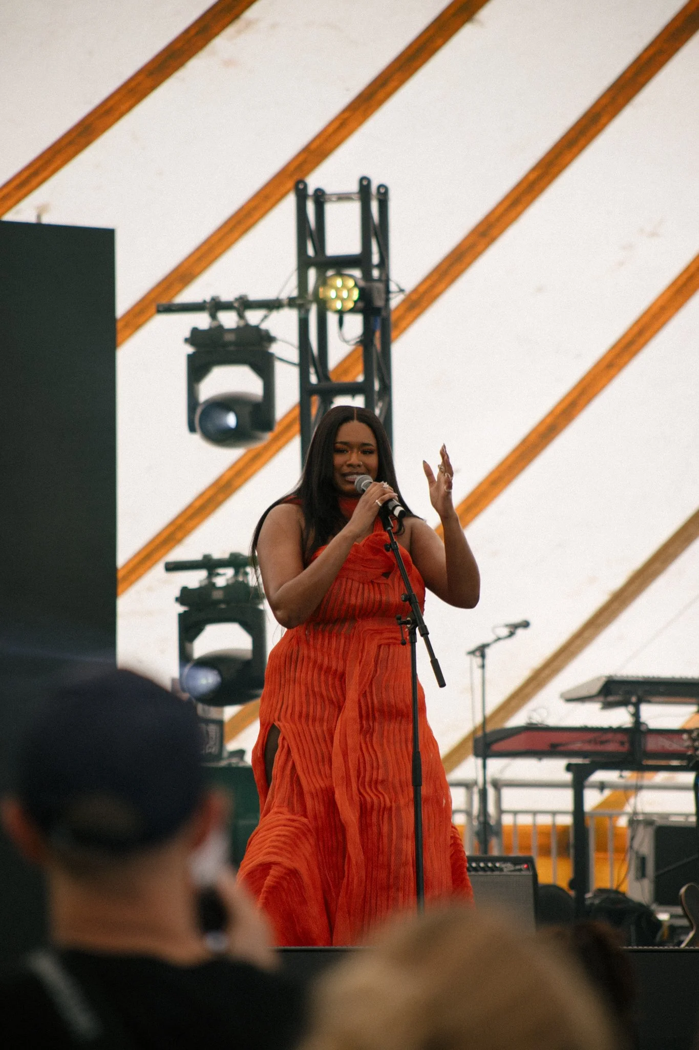 Musician Jamilah Barry in a bright orange dress singing into a microphone on an outdoor stage with musical equipment and wooden beams in the background.