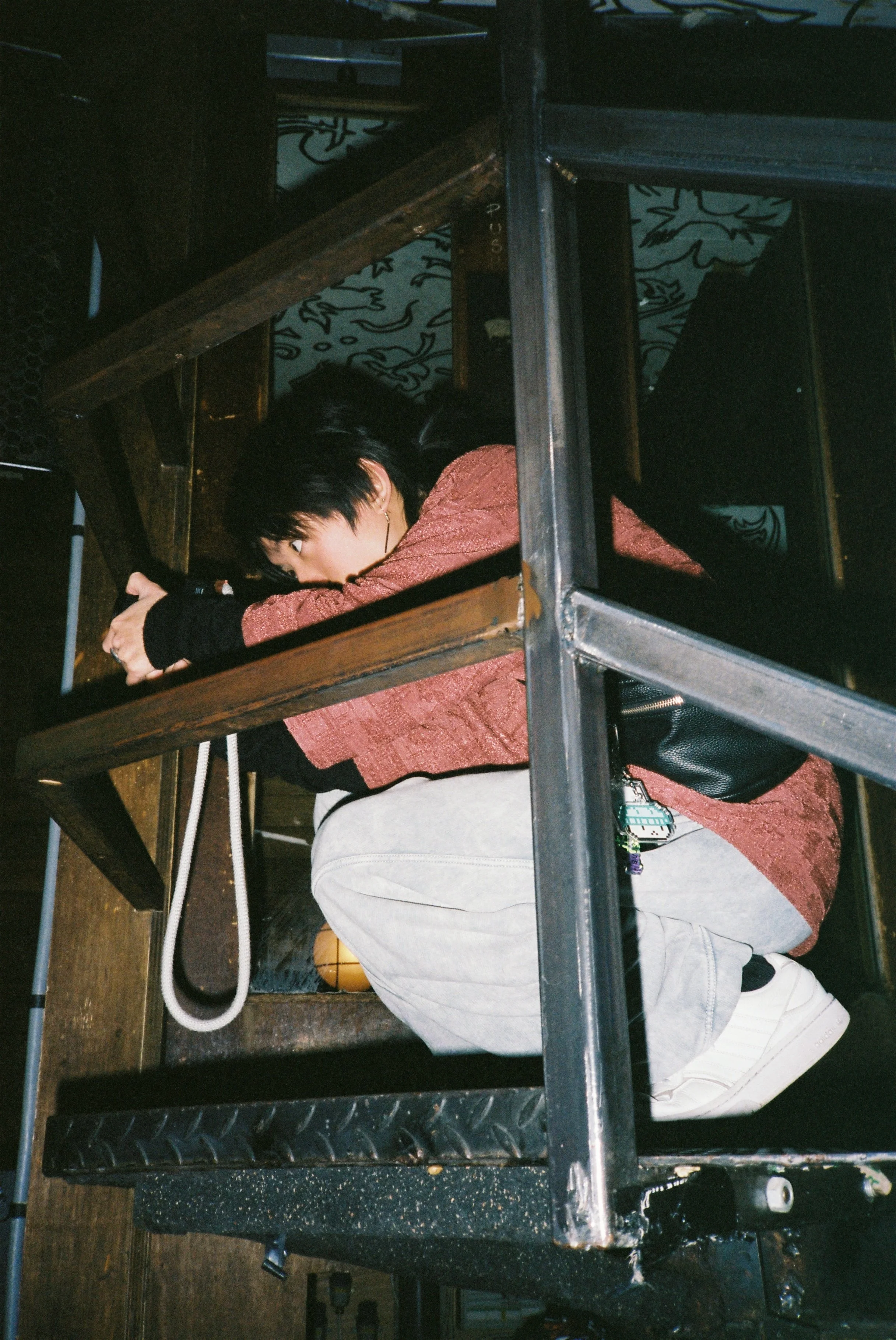 A person with black hair wearing a pink jacket, gray shorts, and white sneakers, lying on the floor under a black metal structure, with arms extended wearing black gloves, holding a camera.