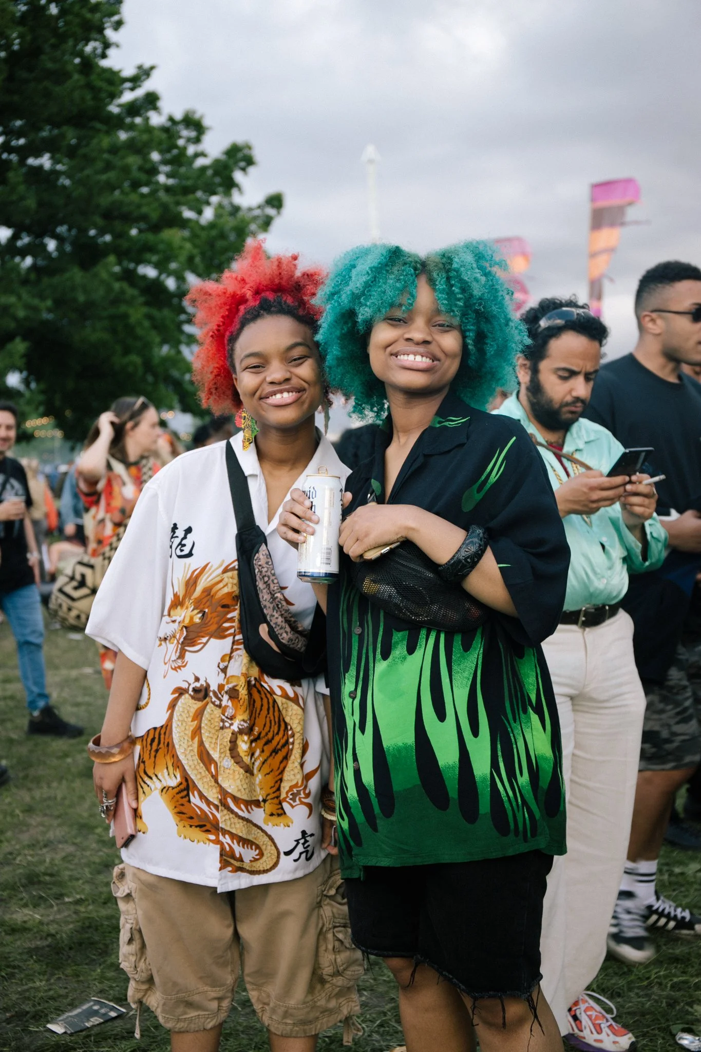 Two young women with colorful curly hair, smiling at a crowded outdoor event, wearing casual and vibrant clothing.