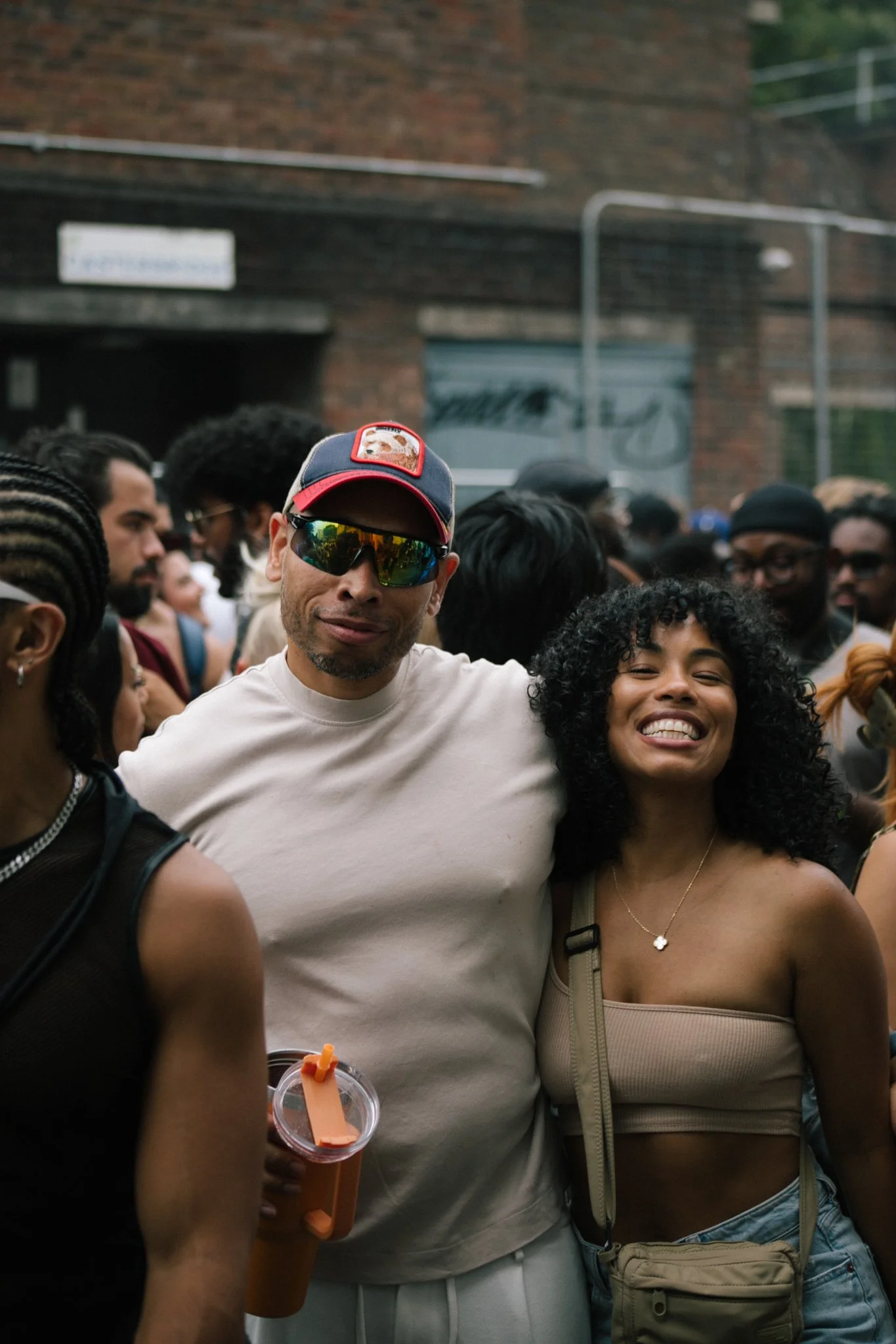 A group of diverse people gathered outdoors, smiling, with some wearing sunglasses. A man in the center wears a cap with a dog patch and reflective sunglasses, and a woman next to him has curly hair and is smiling. The background includes a brick bui