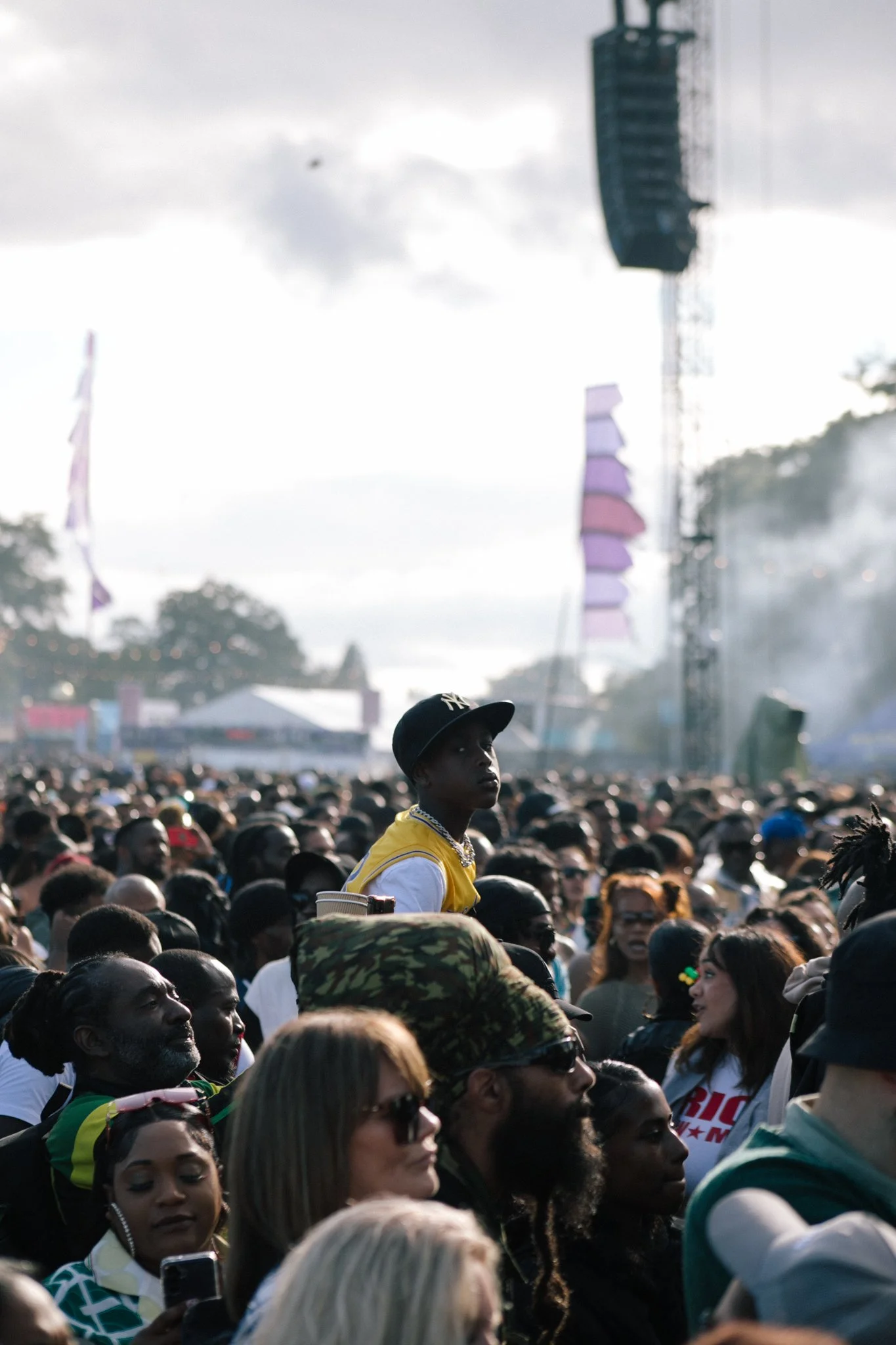 A large crowd gathered outdoors at a festival or concert on a cloudy day, with many people facing toward the stage or focal point, some talking and some focused on their phones.