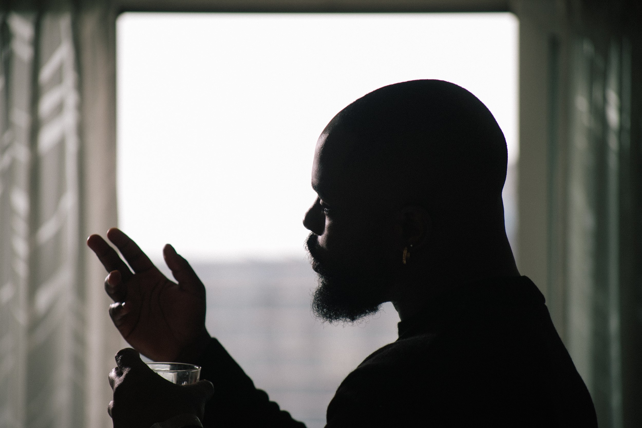 Silhouette of a man with a beard holding a glass in one hand and gesturing with the other, set against a backlit window.