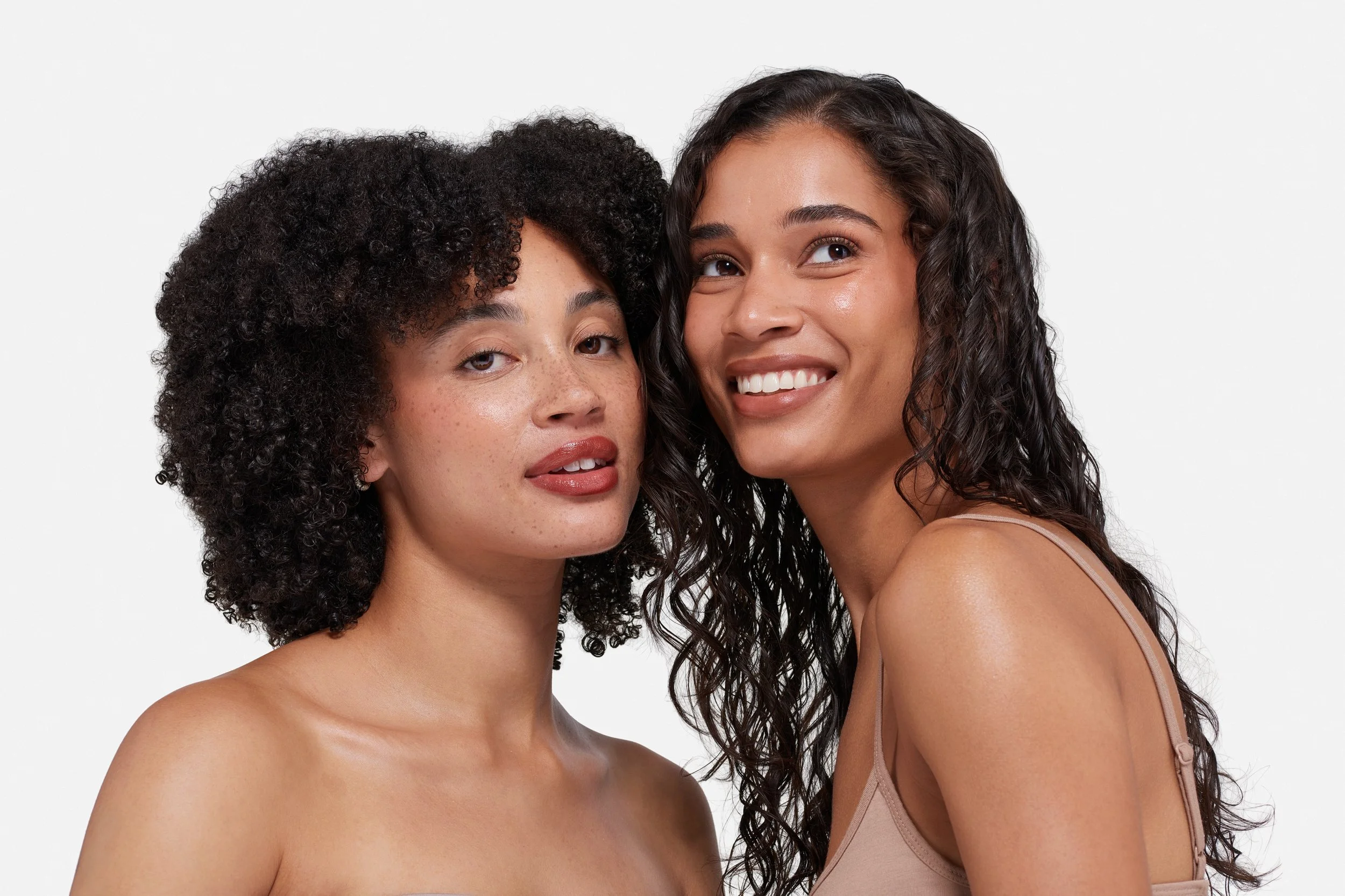 Two young women with curly hair smiling and posing close together against a white background.