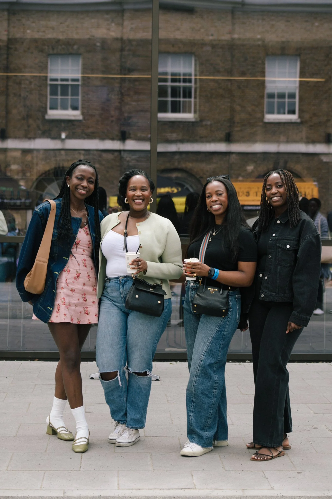 Four women standing together in front of a glass window, smiling and holding drinks on a city street.