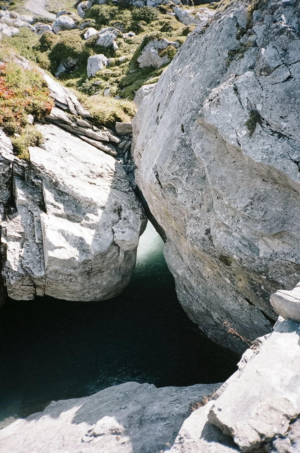 Large rocks and boulders surround a narrow water channel in a rugged natural landscape with some sparse vegetation.