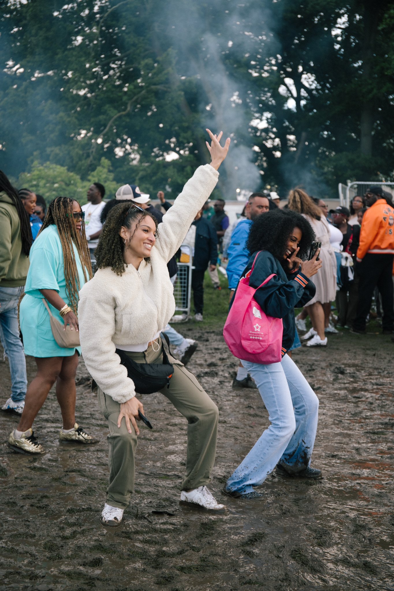 Young woman dancing and smiling with her hand raised during an outdoor event, surrounded by a crowd of people on a muddy field with trees in the background.