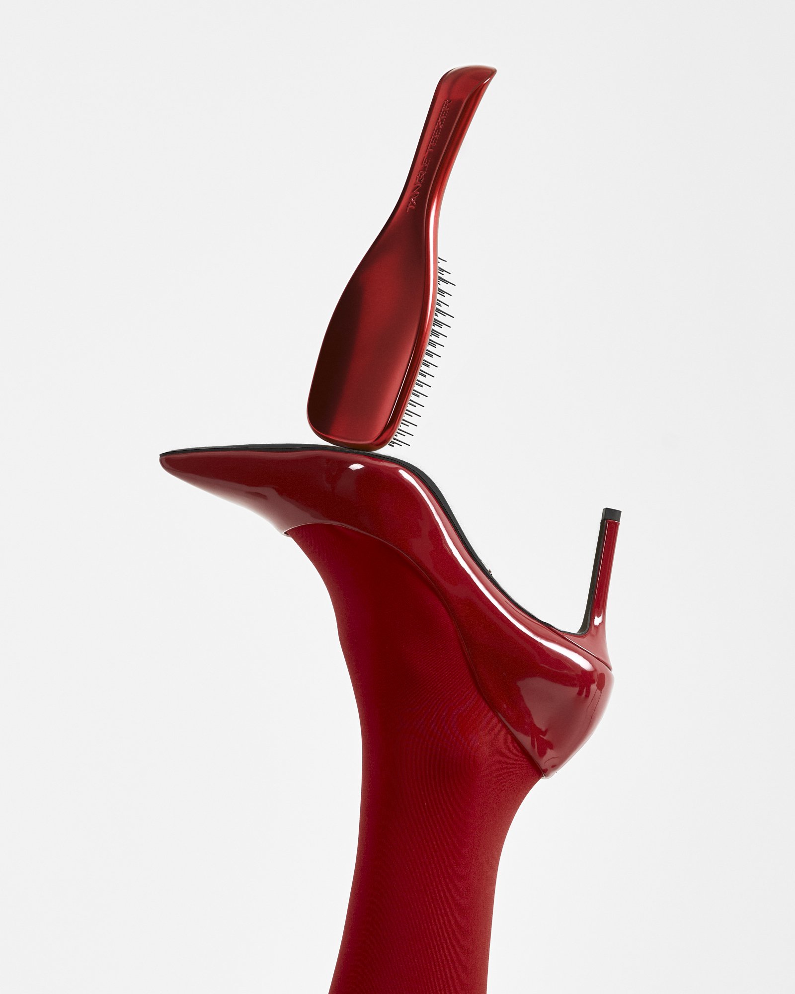 A leg raise upwards, in red tights with wearing a red high-heeled shoe with a red hairbrush poiting upwards from the sole of the heel- seen from a side angle against a plain background.