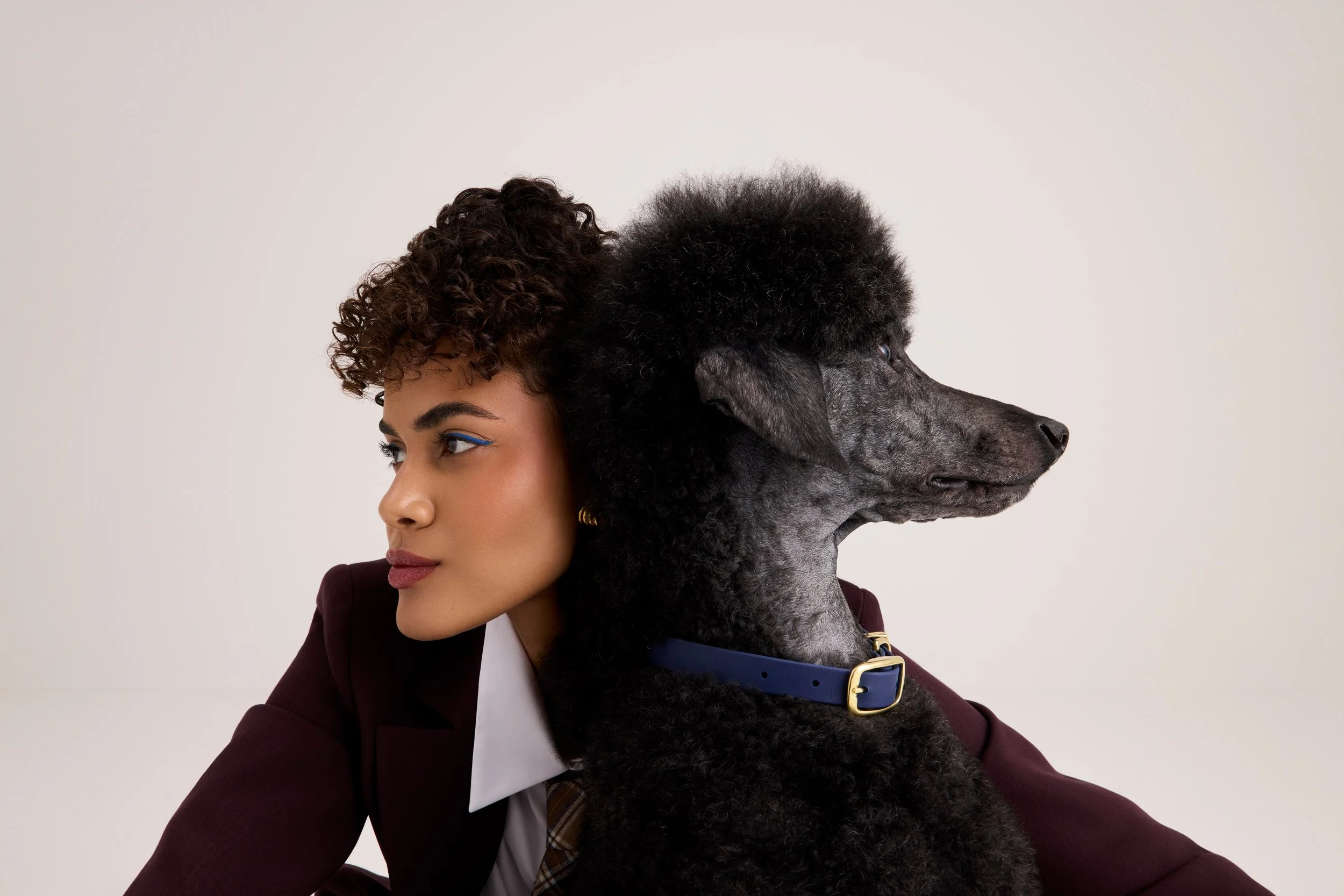 A woman with curly hair, makeup, and a maroon blazer, facing left, with a large black poodle dog with curly fur and a blue collar, facing right, against a plain white background.
