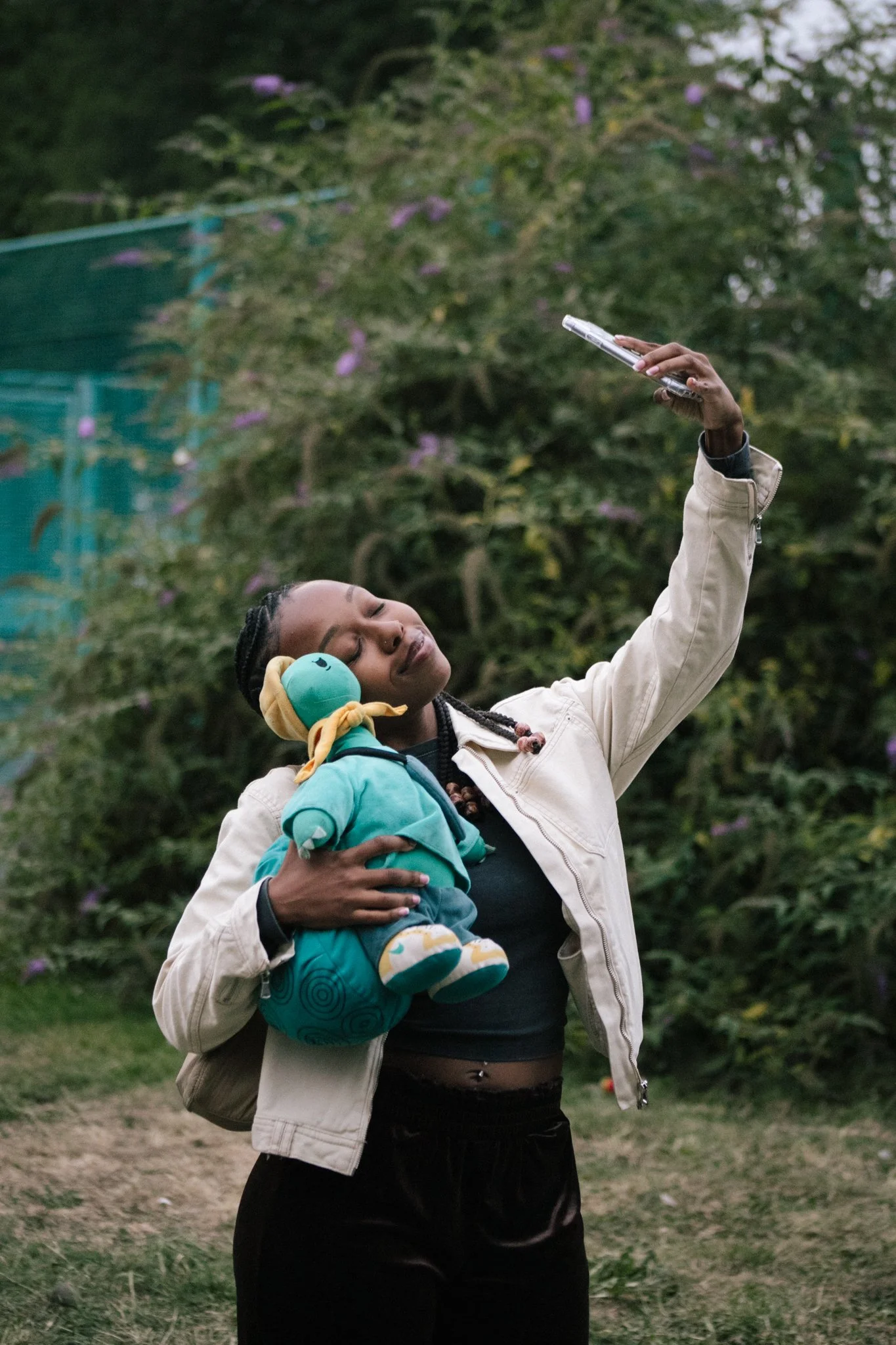 A woman is taking a selfie outdoors, holding a colorful plush turtle toy in her arm. She has braided hair, wears a beige jacket and black pants, and is smiling with eyes closed against a background of greenery and purple flowers.