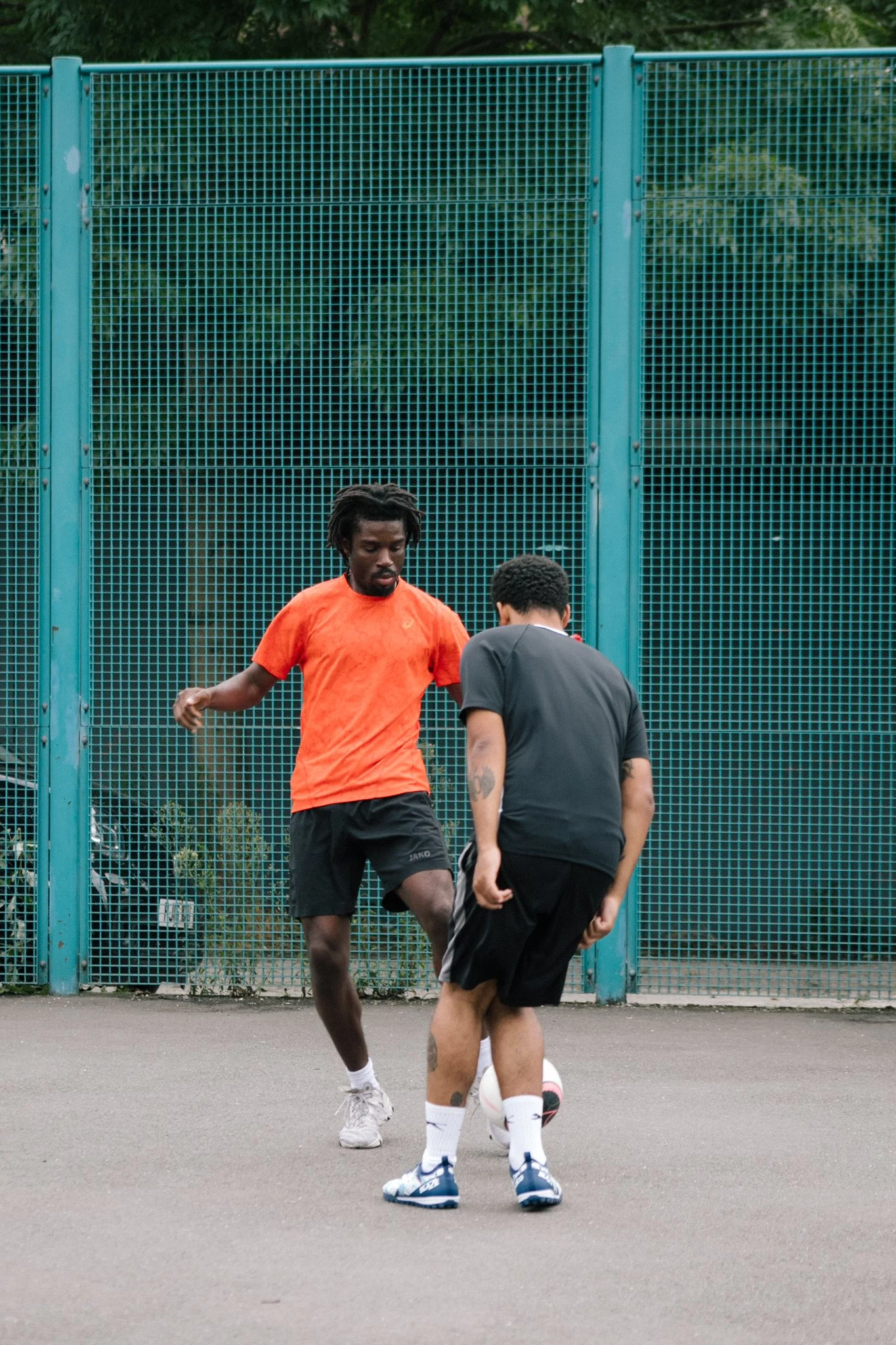Two young men playing football on an outdoor court, with a green chain-link fence in the background.