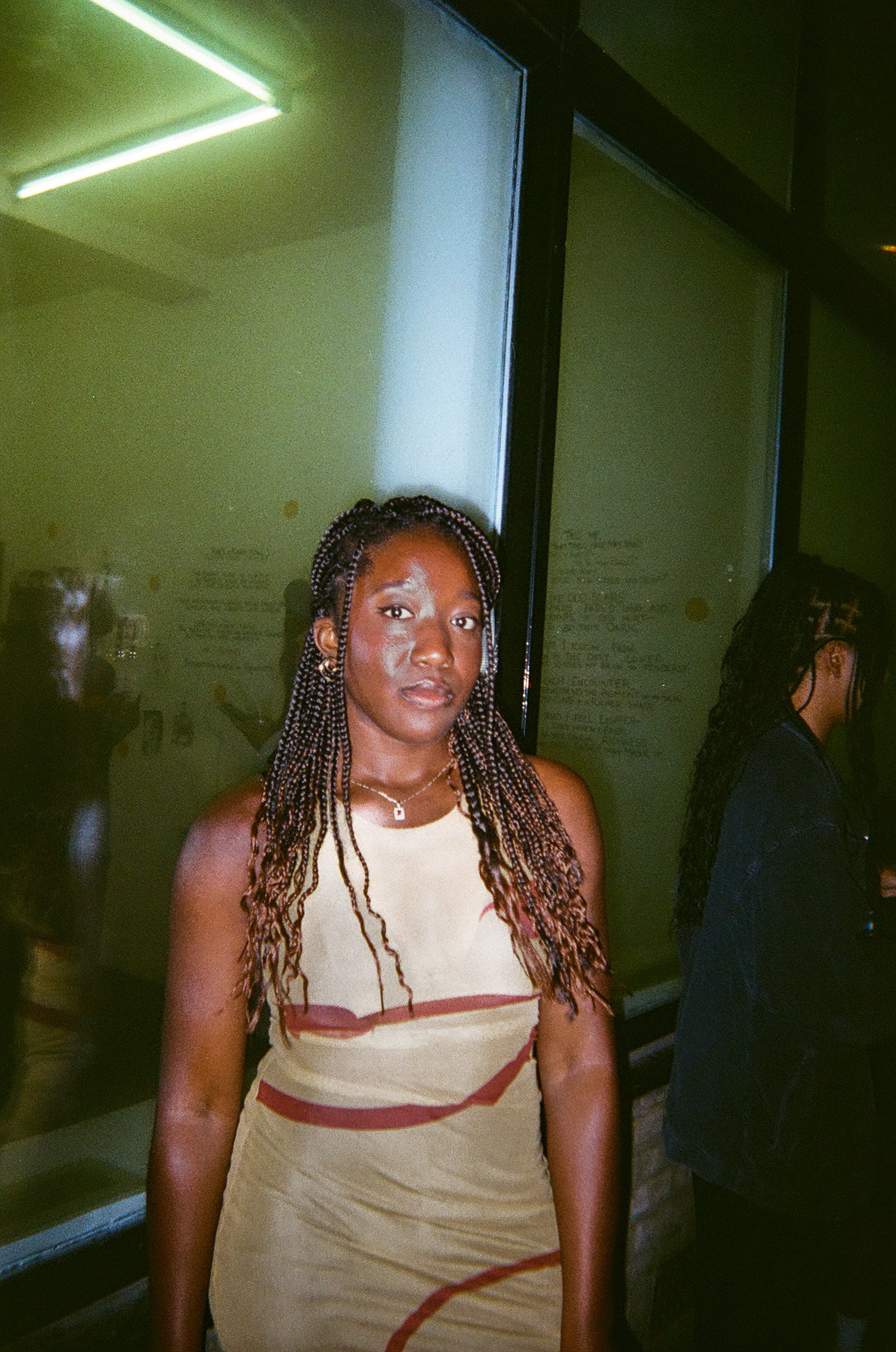 A woman with braided hair wearing a sleeveless dress with a light background and dark markings, standing near a wall with glass panels.