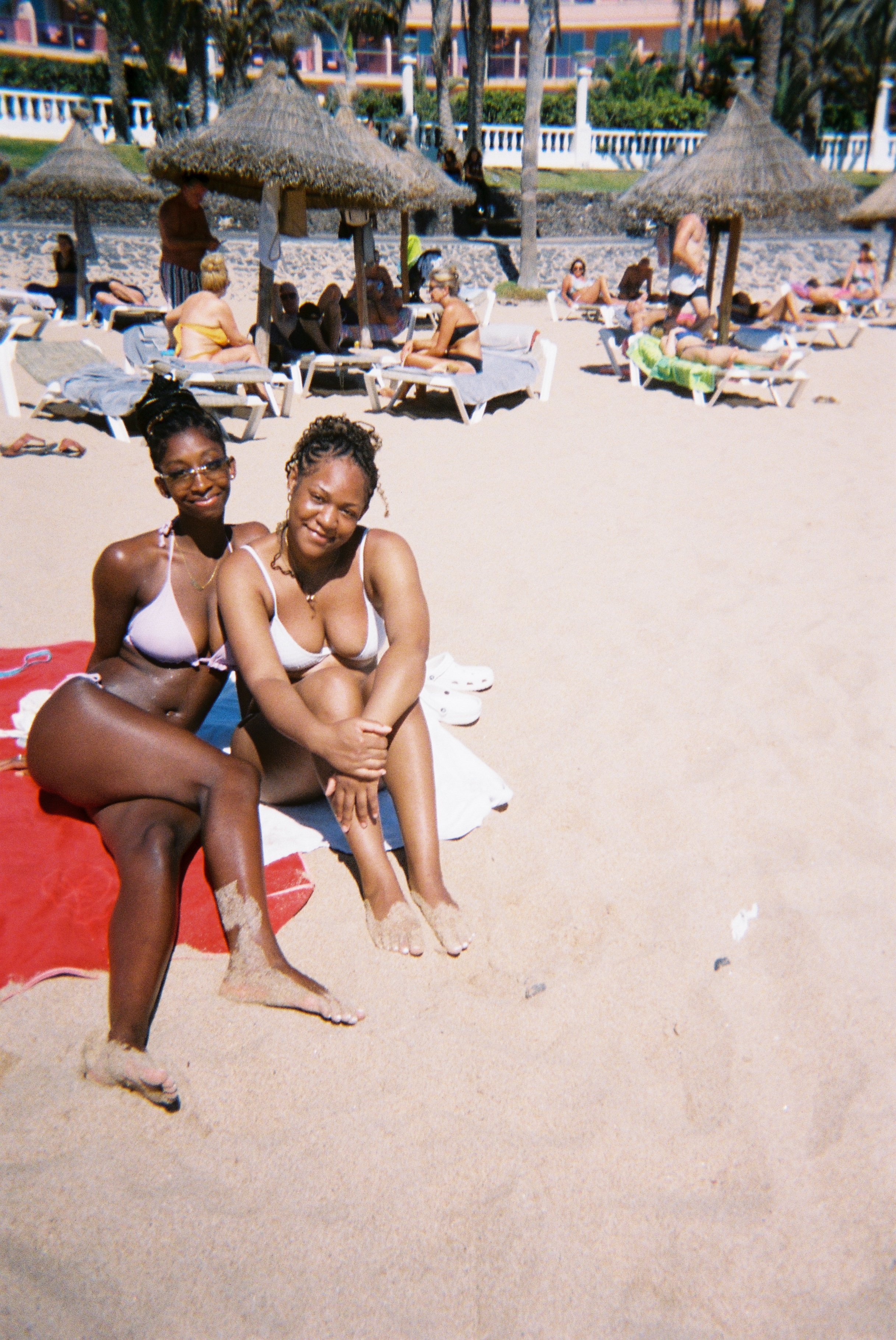 Two women sitting on the sand at a beach, with their feet buried in the sand. They are dressed in swimwear and smiling at the camera, with beach umbrellas and other people relaxing in the background.