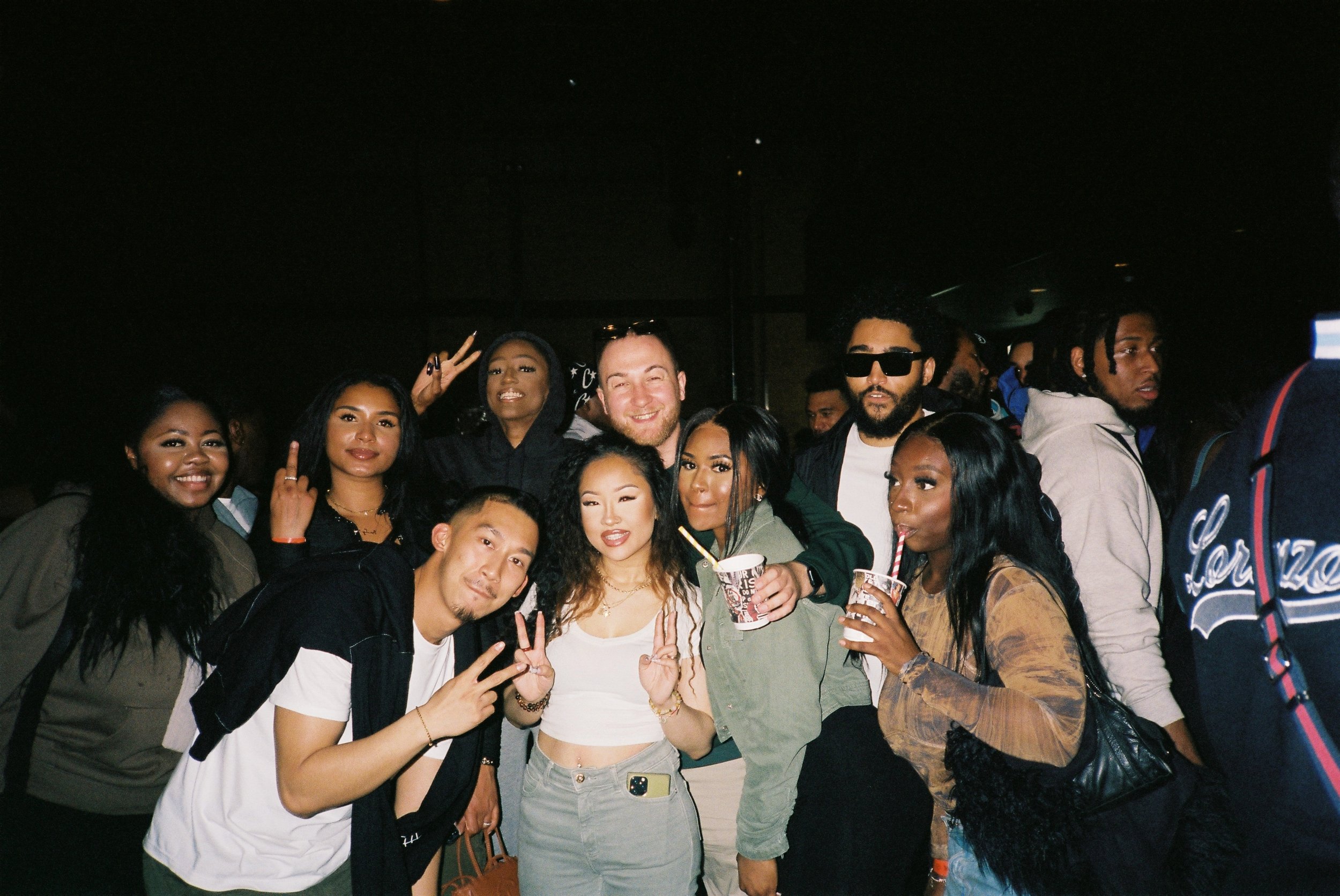 Group of diverse young people at a party, some smiling and making peace signs, holding drinks, in a dimly lit setting.