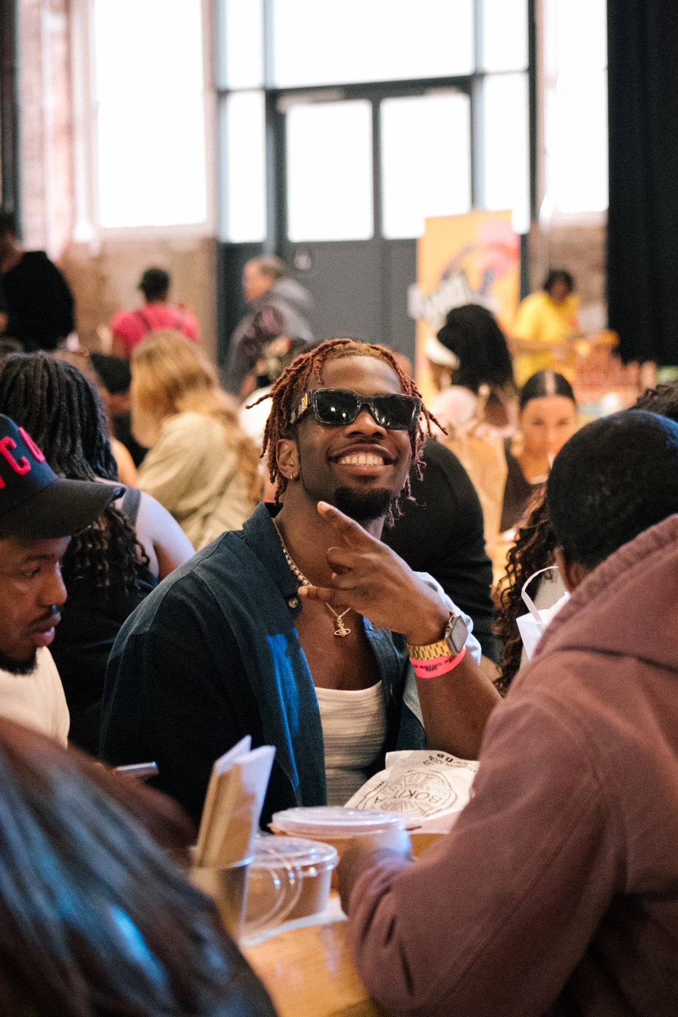A group of people gathered indoors, socializing, with one person in the foreground wearing sunglasses and smiling, surrounded by others at tables with food and drinks.